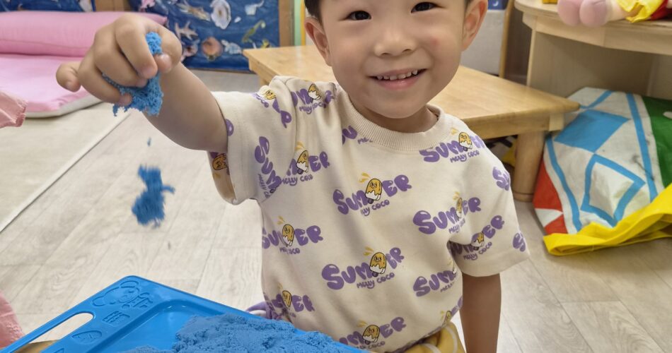 환하게 웃는 지호가 파란색 키네틱 샌드를 한 손에 들고 카메라를 응시하고 있습니다. 테이블 위에는 파란색 트레이에 샌드가 담겨 있고, 뒤로는 교실 풍경이 보입니다.