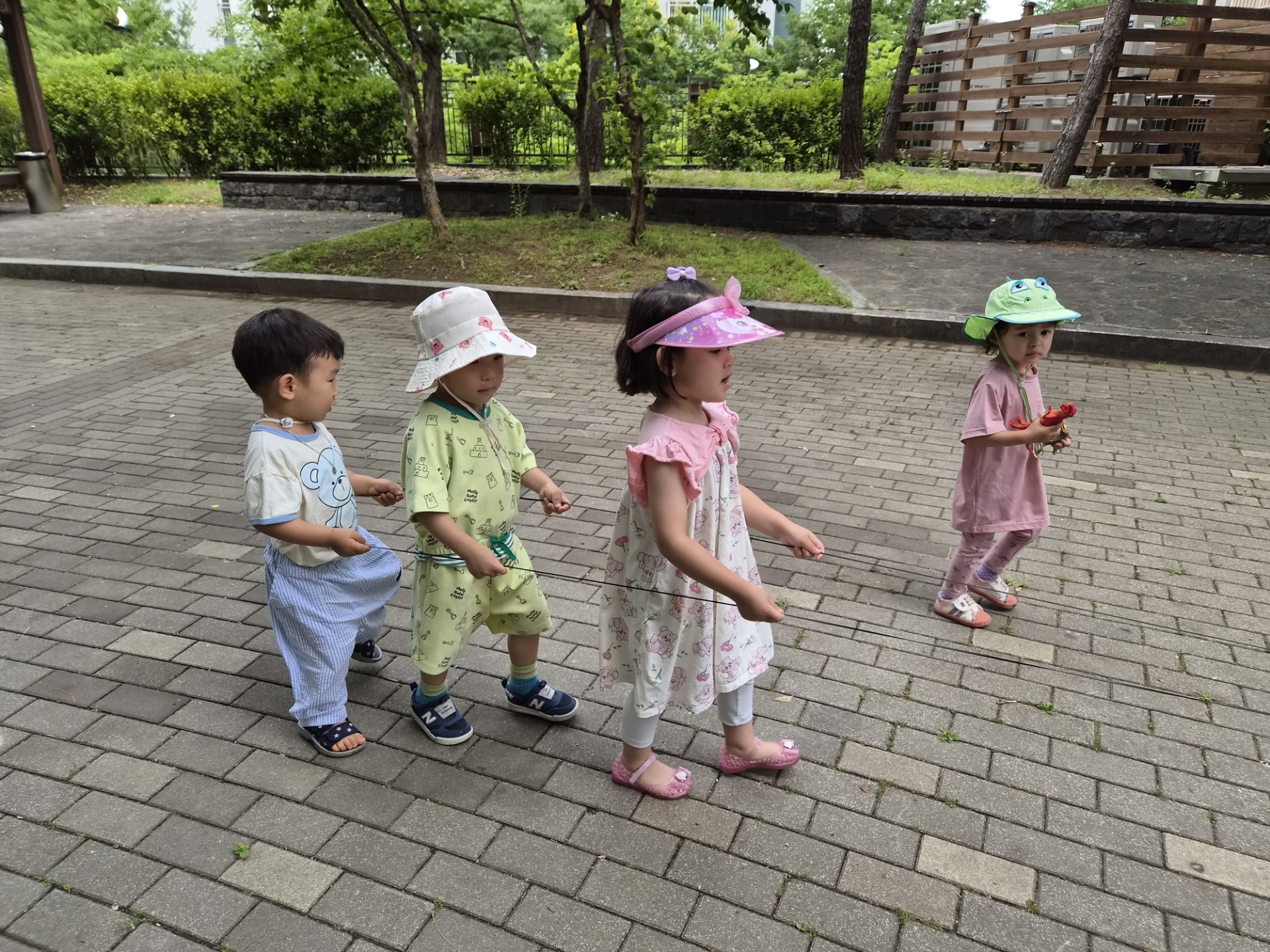 지호가 흰색 줄무늬 바지와 흰색 모자를 쓰고 친구들과 함께 야외에서 일렬로 서서 앞을 보며 걷고 있습니다. 다른 세 명의 친구들도 각자 모자를 쓰고 줄을 잡고 있습니다. 뒤로는 나무와 길이 보입니다.