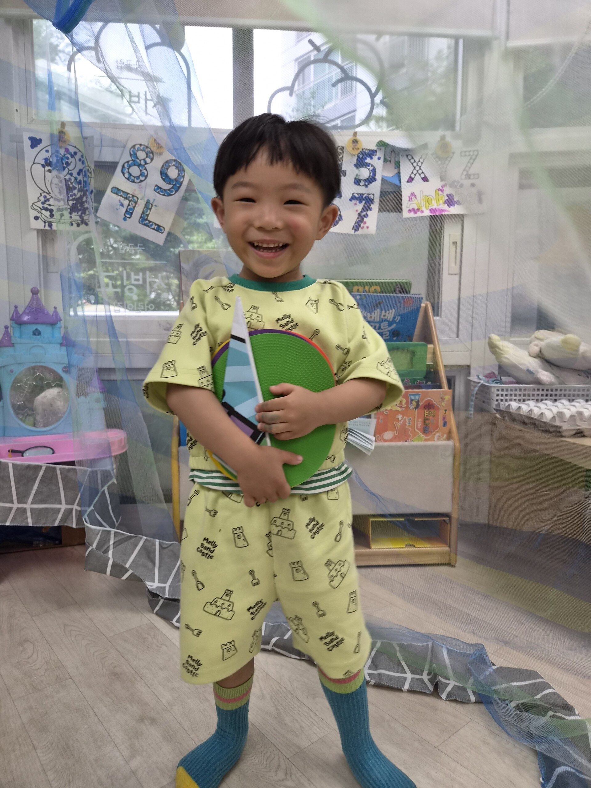 밝은 연두색 옷을 입은 지호가 여러 색깔의 원형 놀잇감을 양손으로 안고 서서 환하게 웃고 있습니다. 배경은 창문과 책장, 그리고 천장에 드리워진 푸른 천으로 꾸며진 교실입니다.