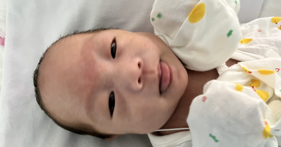 흰 침대 시트 위에 누워있는 신생아 지호가 노란 오리 무늬 옷을 입고 정면을 바라보며 혀를 낼름거리고 있다.