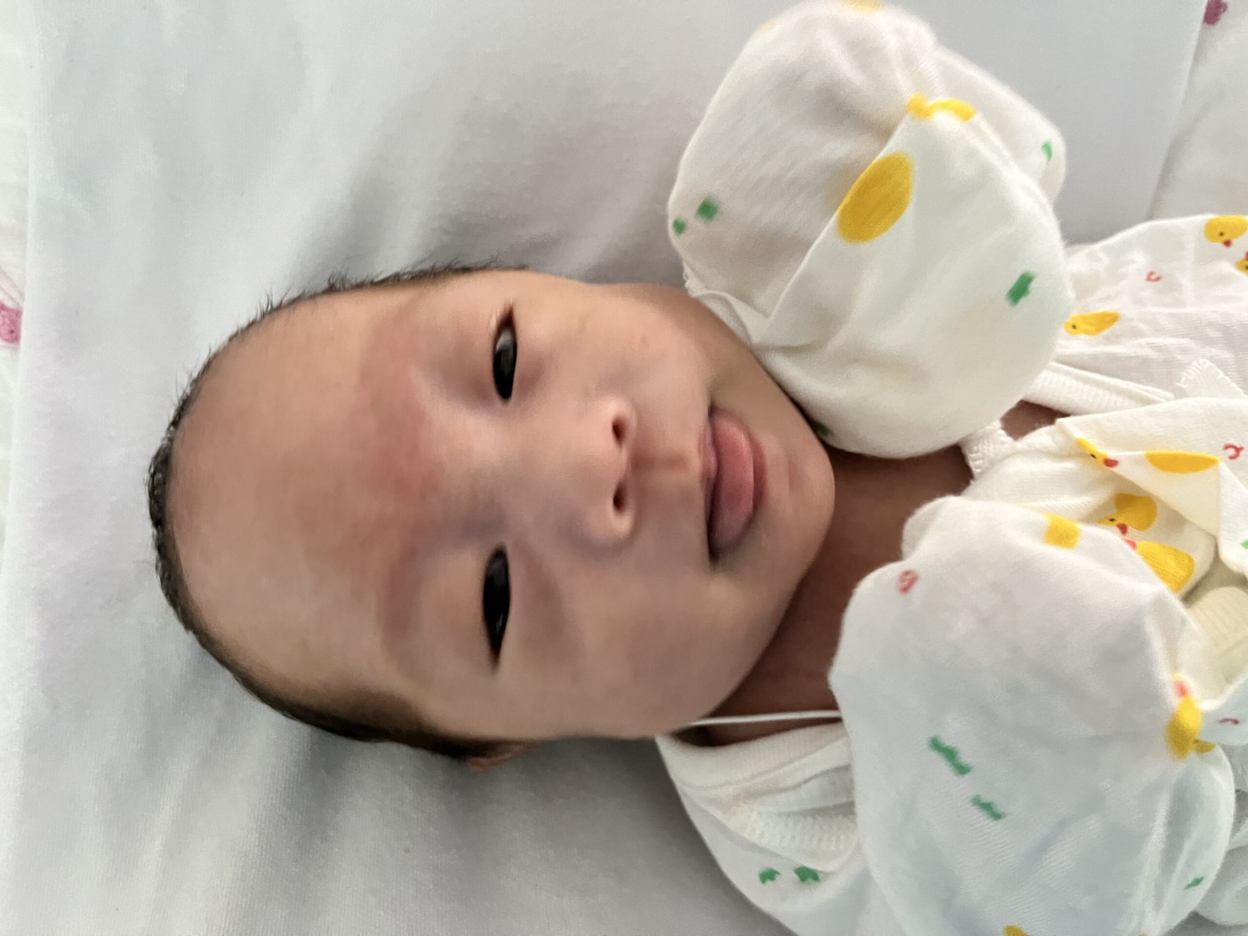 흰 침대 시트 위에 누워있는 신생아 지호가 노란 오리 무늬 옷을 입고 정면을 바라보며 혀를 낼름거리고 있다.