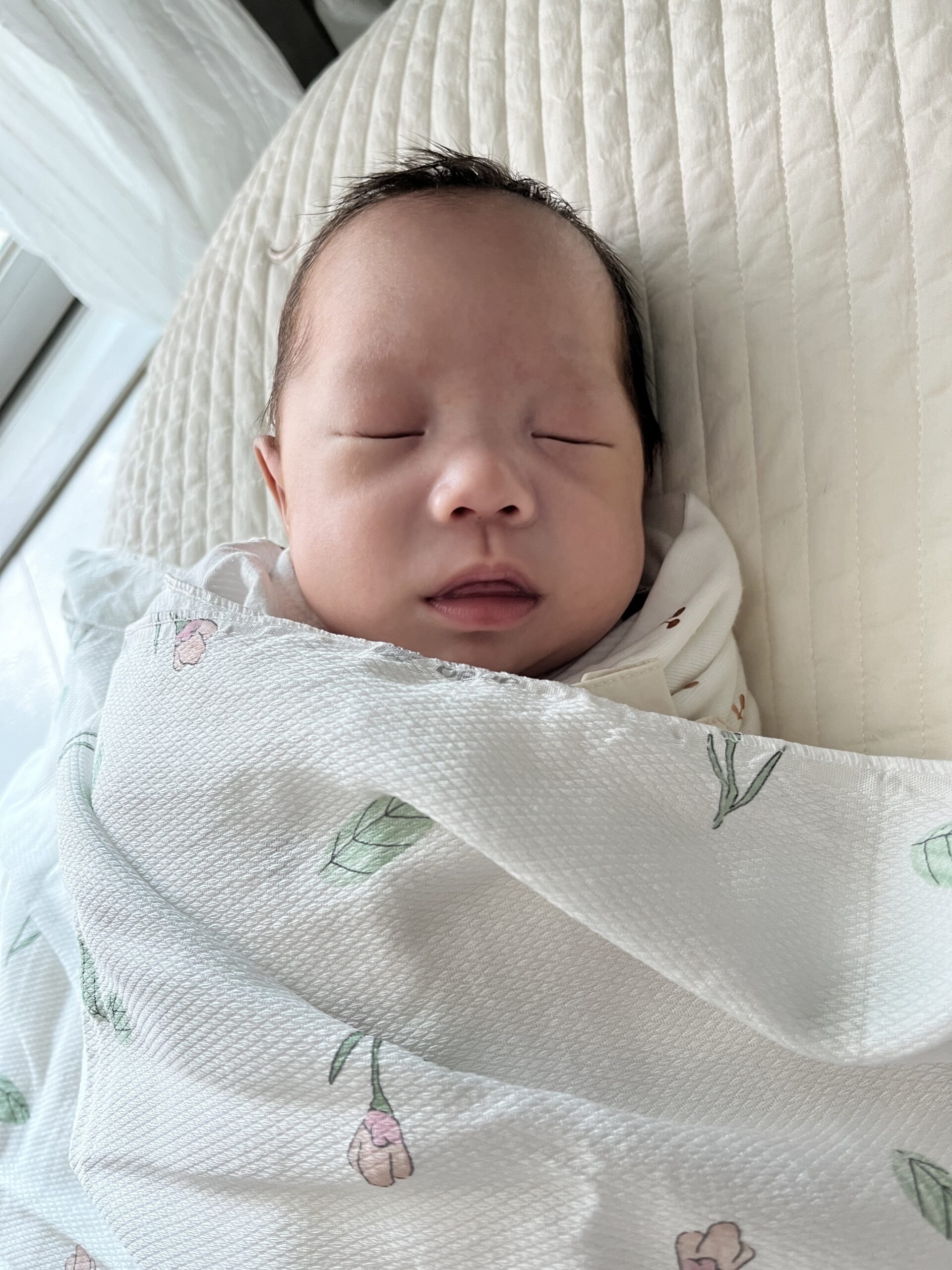 흰색 아기 침대 위에 흰색 바탕에 연두색 잎사귀와 분홍색 꽃무늬가 그려진 블랭킷에 싸여 곤히 잠든 아기 지호의 모습. 머리는 약간 검은색이고 눈은 감겨 있으며 입은 살짝 벌어져 있다.