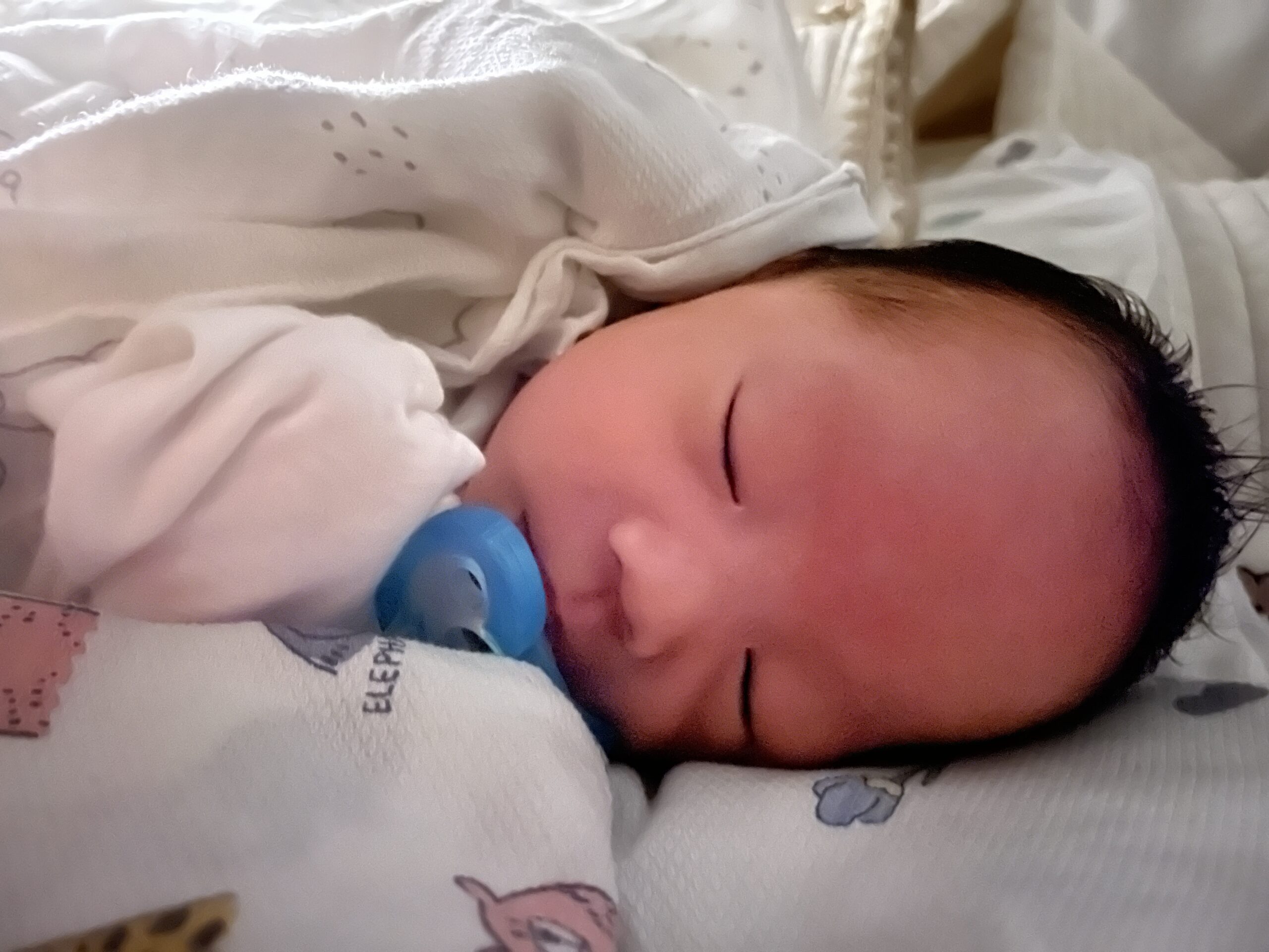 흰색 이불을 덮고 잠든 신생아 지호의 측면 모습. 입에는 파란색 공갈 젖꼭지가 물려 있고 얼굴은 살짝 붉다.