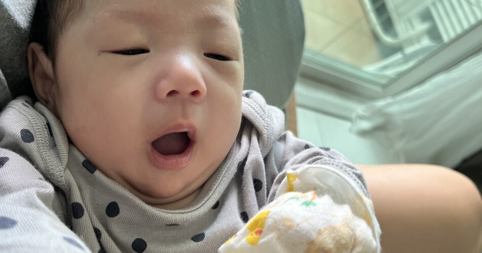 엄마 품에 안겨 회색 도트 무늬 우주복을 입고 입을 크게 벌려 하품하는 아기 지호의 근접 사진. 한 손에는 작은 흰색 벙어리 장갑을 끼고 있다.
