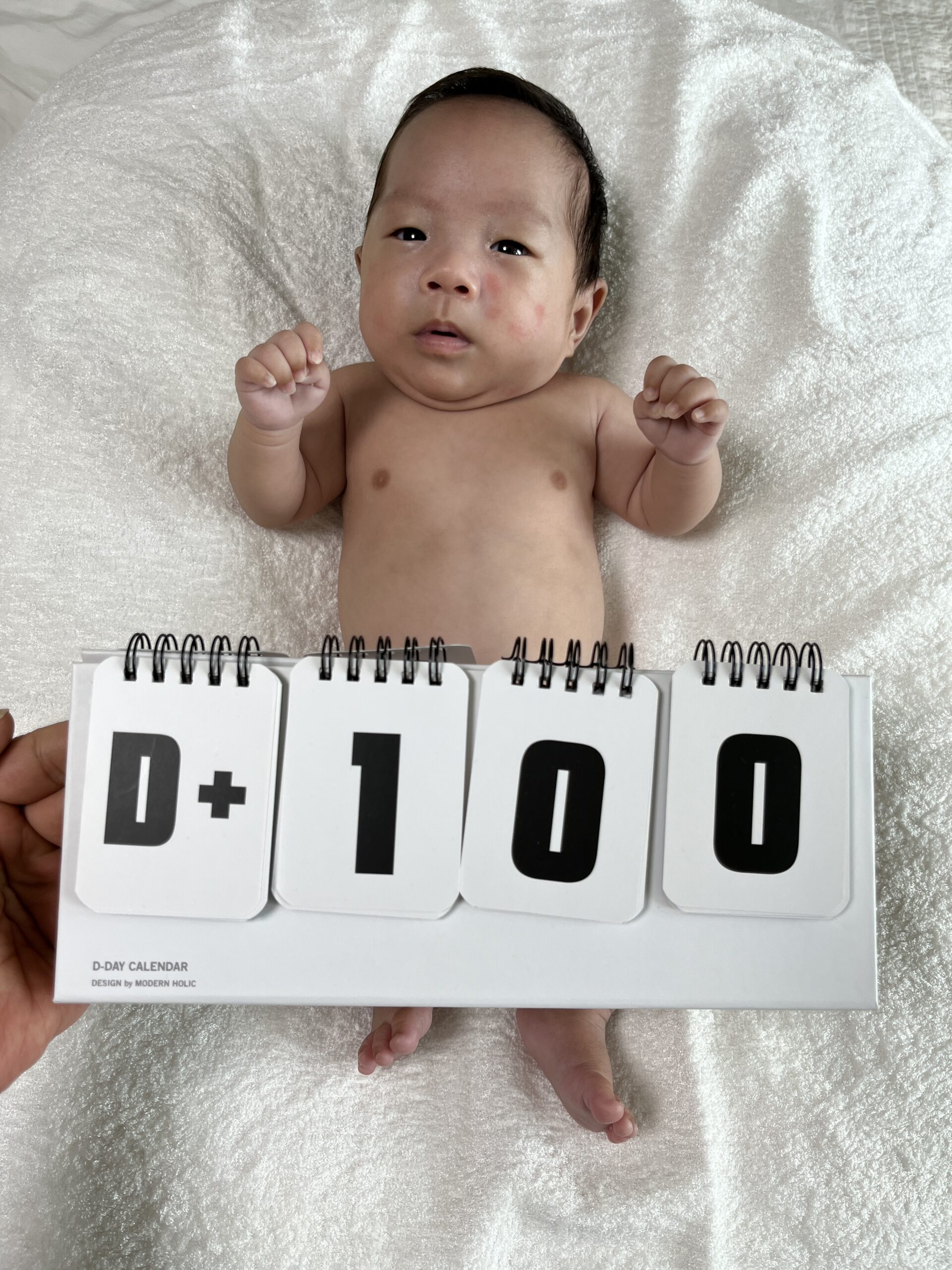 흰색 이불 위에 누워있는 아기 지호의 상체와 다리, 그리고 발 아래 놓인 'D+100'이 표시된 D-DAY 달력. 지호는 카메라를 향해 맑은 눈으로 응시하고 있다.