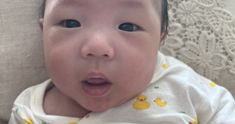 흰색 레이스 베개에 누워 카메라를 응시하는 아기 지호의 클로즈업 사진. 노란 오리 무늬가 있는 흰색 긴팔 우주복을 입고 있으며, 눈을 크게 뜨고 입을 살짝 벌린 채 호기심 어린 표정을 짓고 있다.