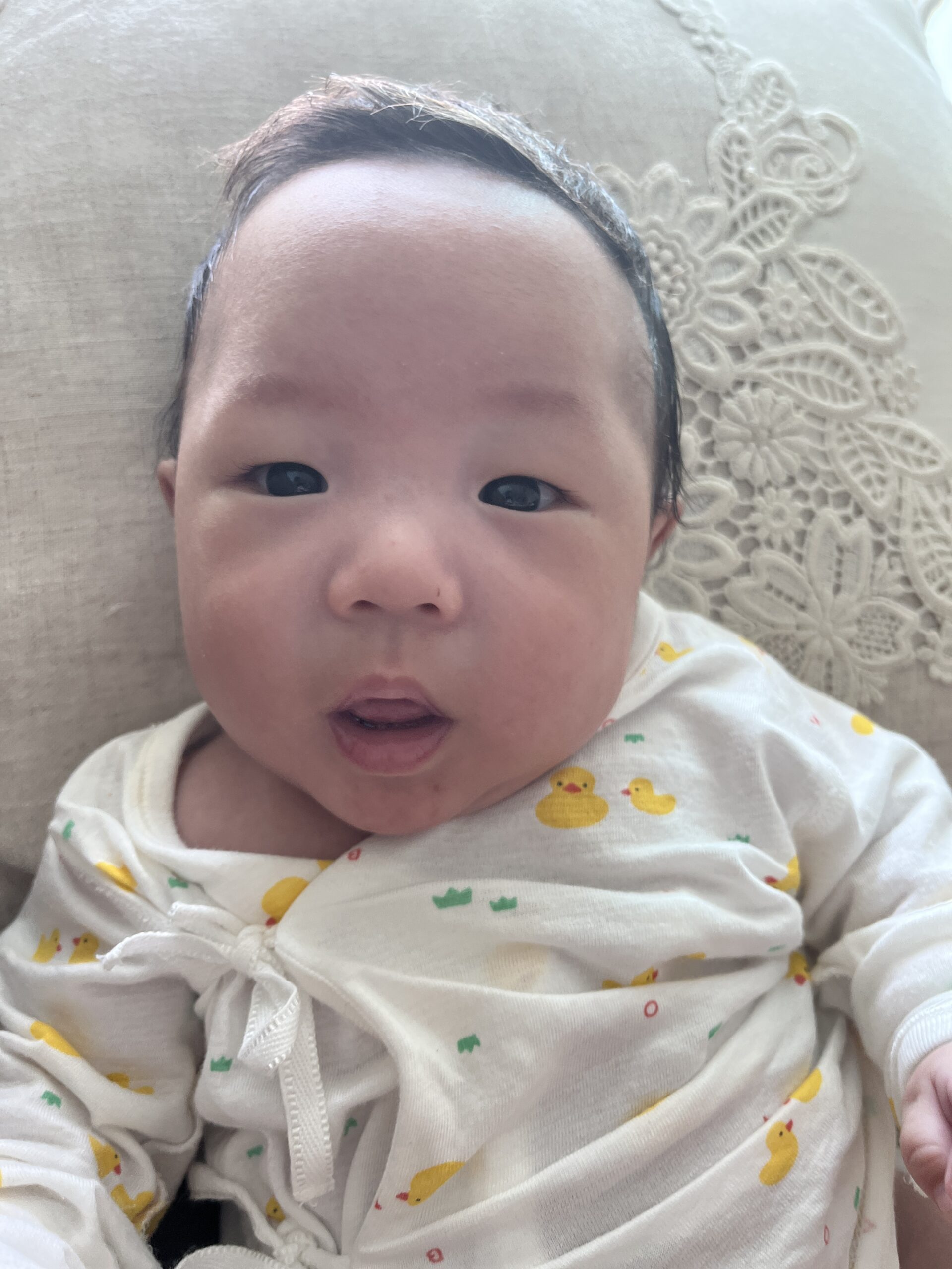 흰색 레이스 베개에 누워 카메라를 응시하는 아기 지호의 클로즈업 사진. 노란 오리 무늬가 있는 흰색 긴팔 우주복을 입고 있으며, 눈을 크게 뜨고 입을 살짝 벌린 채 호기심 어린 표정을 짓고 있다.