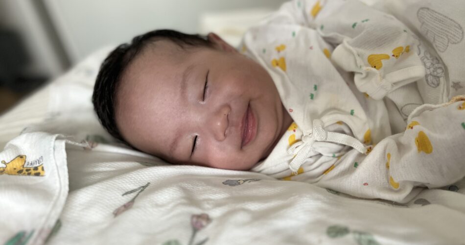 하얀색 병아리 패턴 애착 이불 위에 누워 평화롭게 잠든 아기가 살짝 미소를 짓고 있다. 흰색에 노란색 병아리 무늬가 있는 아기 옷을 입고 있다.