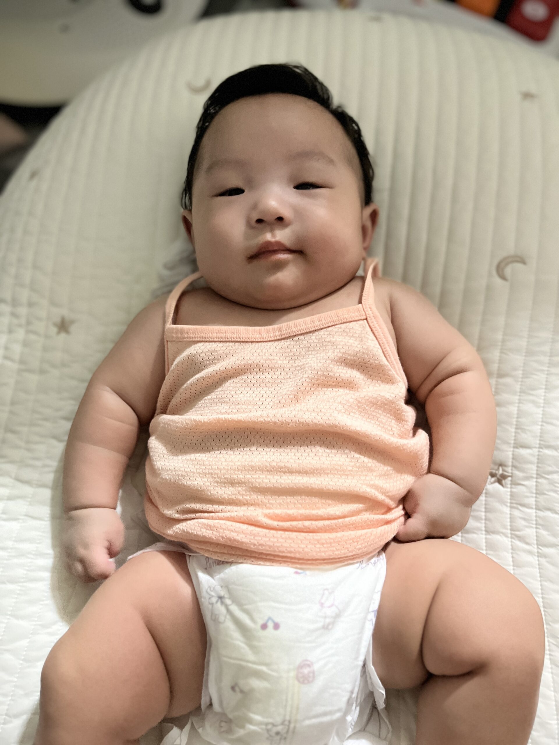 흰색 누빔 아기 매트 위에 누워 카메라를 응시하고 있는 아기 지호의 상반신 모습. 지호는 연한 오렌지색 민소매 상의와 기저귀를 착용하고 있으며, 통통한 팔과 허벅지가 눈에 띈다. 어둡고 윤기 있는 머리카락은 단정하게 뒤로 넘어가 있다.