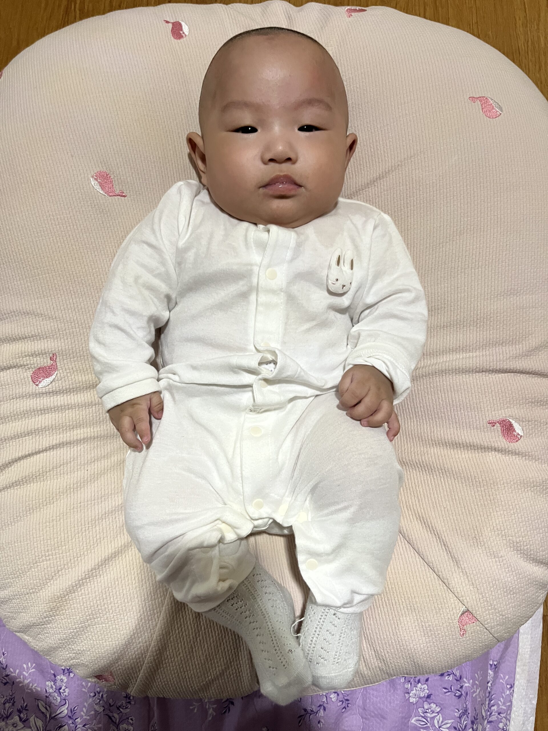 분홍색 토끼 패턴 아기 매트 위에 누워 카메라를 똑바로 바라보고 있는 아기 지호. 흰색 토끼 그림이 있는 긴팔 상하복과 흰색 양말을 신고 있다.
