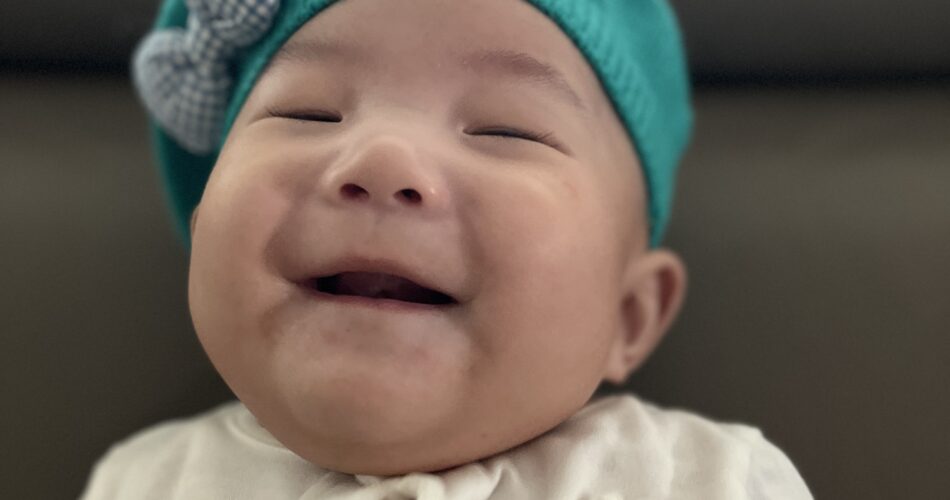 터키색 베레모를 쓰고 눈을 감고 활짝 웃고 있는 아기 지호의 상반신 클로즈업 사진. 흰색 긴팔 상의에는 토끼 자수가 새겨져 있다.