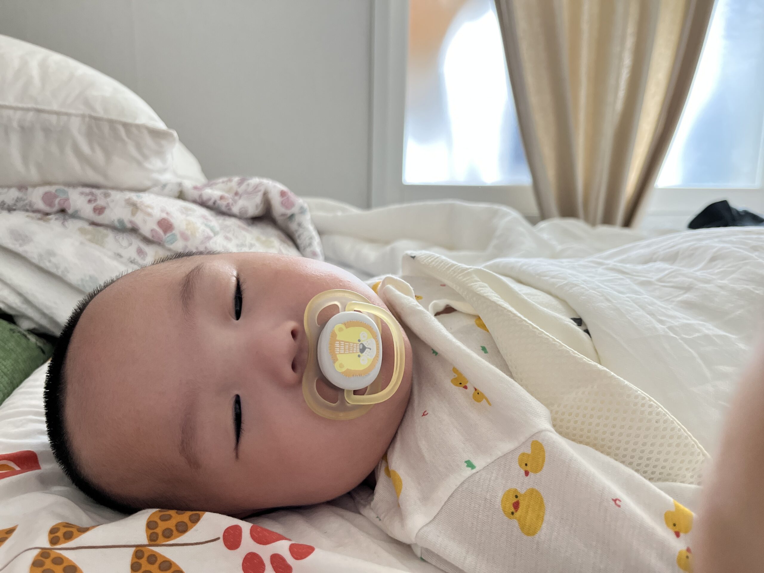 창가 옆 침대 위에서 노란색 오리 모양 쪽쪽이를 물고 편안하게 잠든 아기 지호. 노란색 병아리 패턴의 흰색 잠옷을 입고 있다.