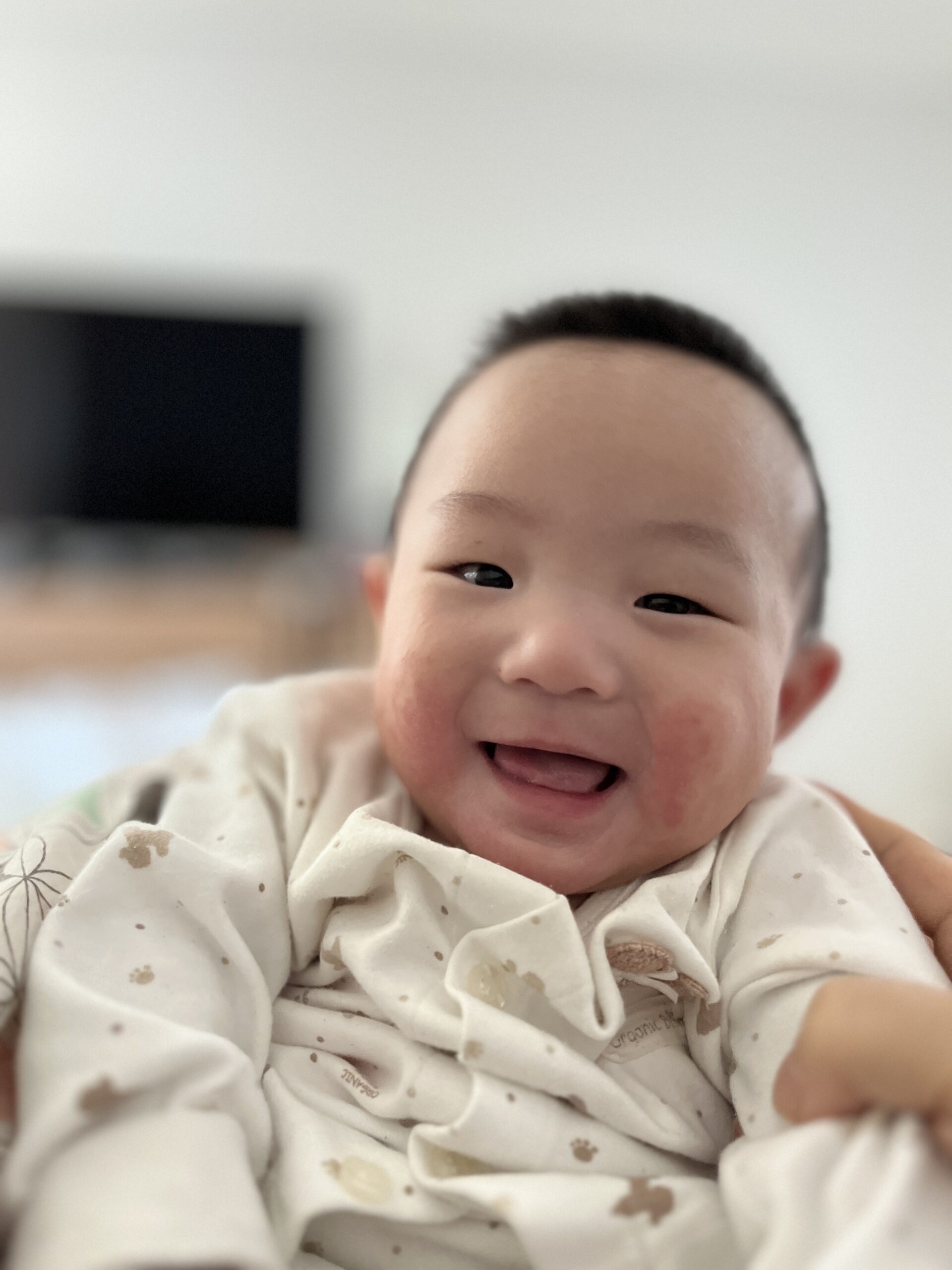 아기가 카메라를 향해 환하게 웃고 있다. 볼은 붉게 상기되어 있고, 해맑은 눈빛으로 정면을 바라본다.