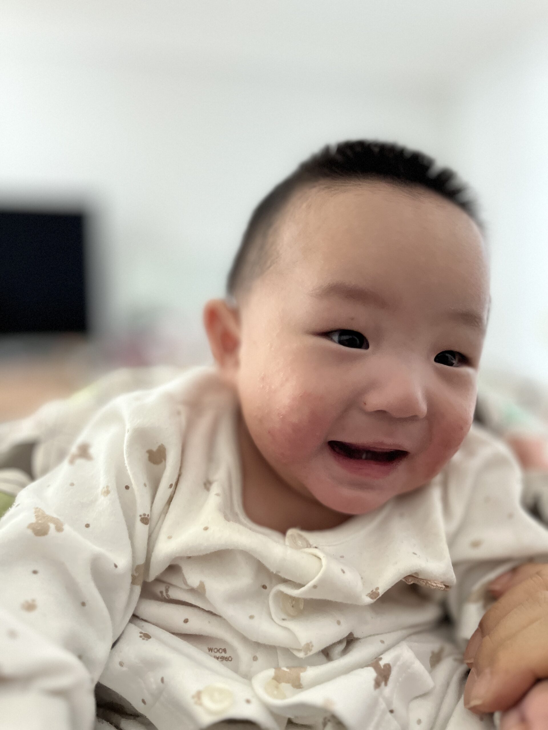 흰색 무늬 옷을 입은 아기 지호가 옆을 보며 환하게 웃고 있는 모습. 얼굴에 약간 붉은 기가 올라와 있다.