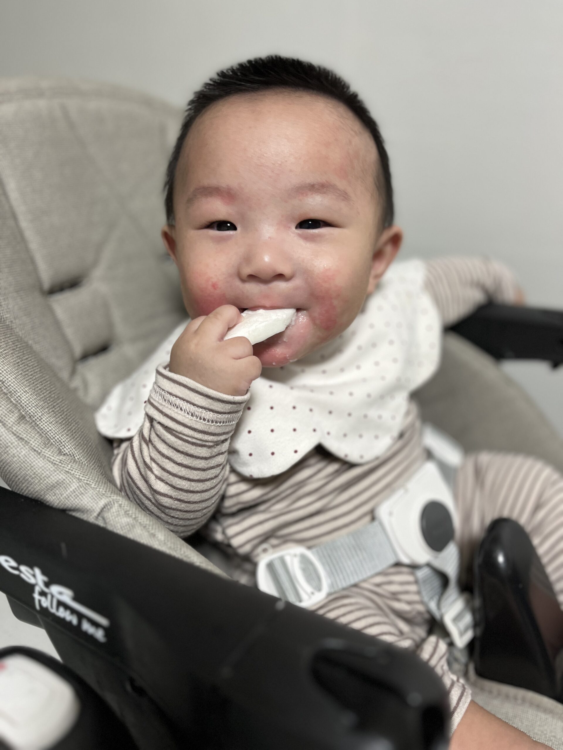 줄무늬 우주복과 물방울무늬 턱받이를 한 아기가 회색 아기 의자에 앉아 한 손에 간식을 들고 먹으며 카메라를 보고 환하게 웃고 있다. 얼굴에 붉은 기가 살짝 보인다.