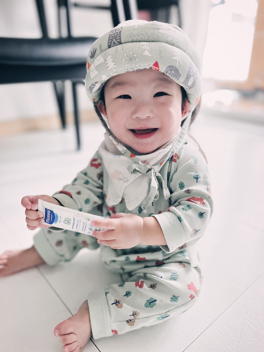 연두색 무늬 옷을 입고 보호 헬멧을 쓴 아기 지호가 바닥에 앉아 카메라를 향해 환하게 웃고 있다. 손에는 작은 로션 튜브를 들고 있다.