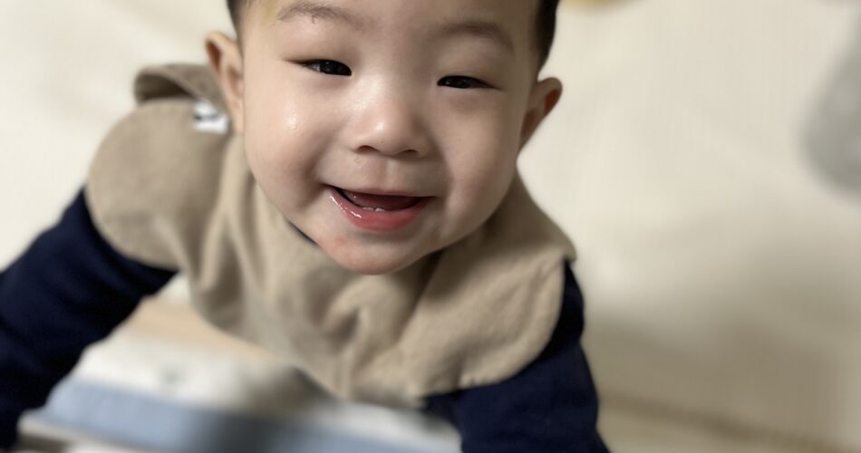 폭신한 침대 위에서 몸을 일으켜 카메라를 향해 활짝 웃고 있는 아기 지호의 모습. 베이지색 턱받이와 남색 상의를 입고 있다.