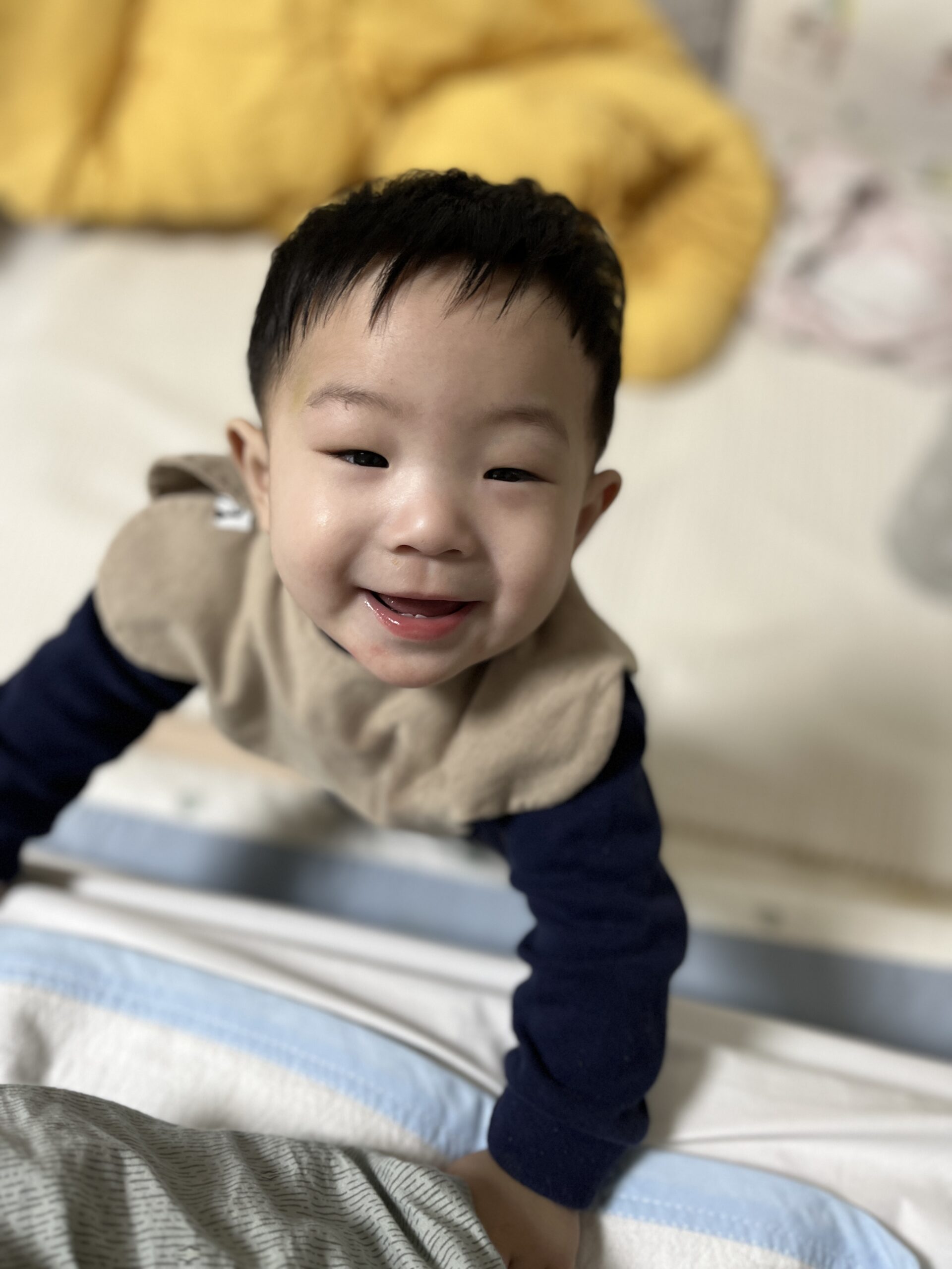 폭신한 침대 위에서 몸을 일으켜 카메라를 향해 활짝 웃고 있는 아기 지호의 모습. 베이지색 턱받이와 남색 상의를 입고 있다.