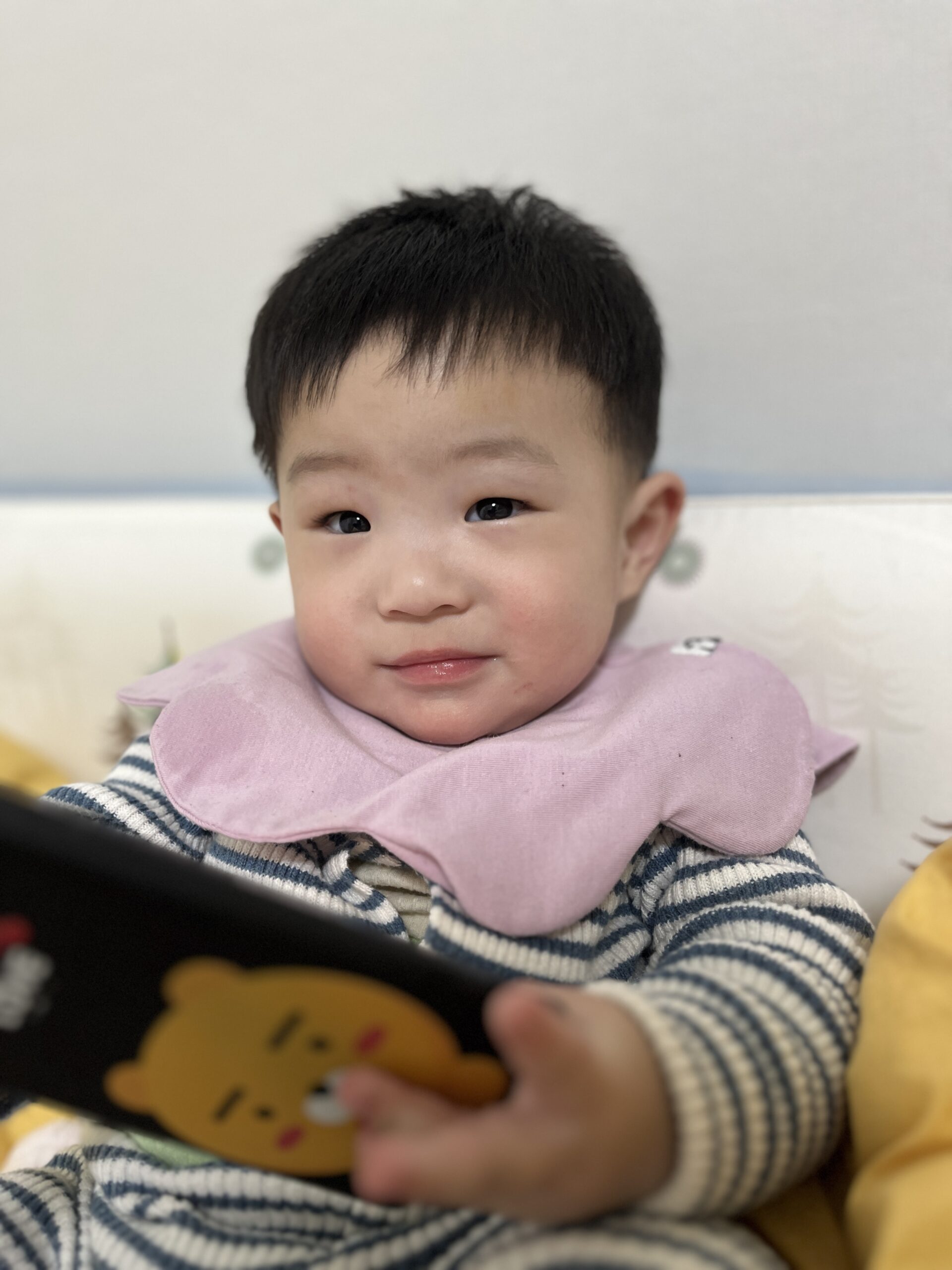 분홍색 턱받이와 줄무늬 상의를 입은 아기 지호가 정면을 보며 무언가를 응시하고 있다. 한 손에 노란 캐릭터가 그려진 검은색 기기를 들고 있다.