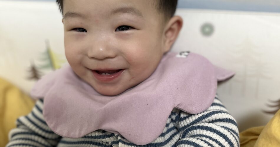 스트라이프 무늬 상의와 핑크색 스카프빕을 한 지호가 휴대폰을 들고 침대에 앉아 환하게 웃고 있는 모습.