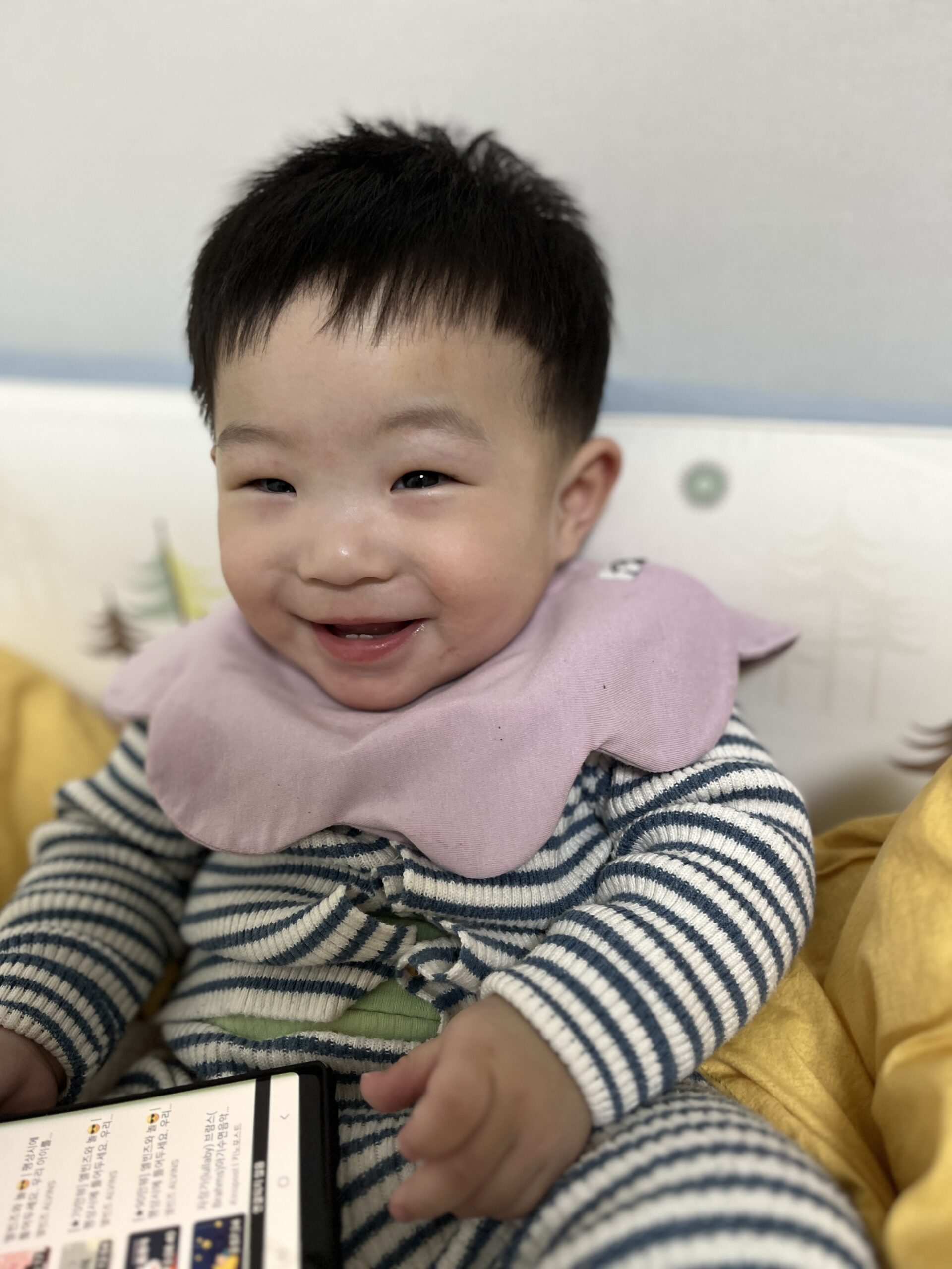 스트라이프 무늬 상의와 핑크색 스카프빕을 한 지호가 휴대폰을 들고 침대에 앉아 환하게 웃고 있는 모습.