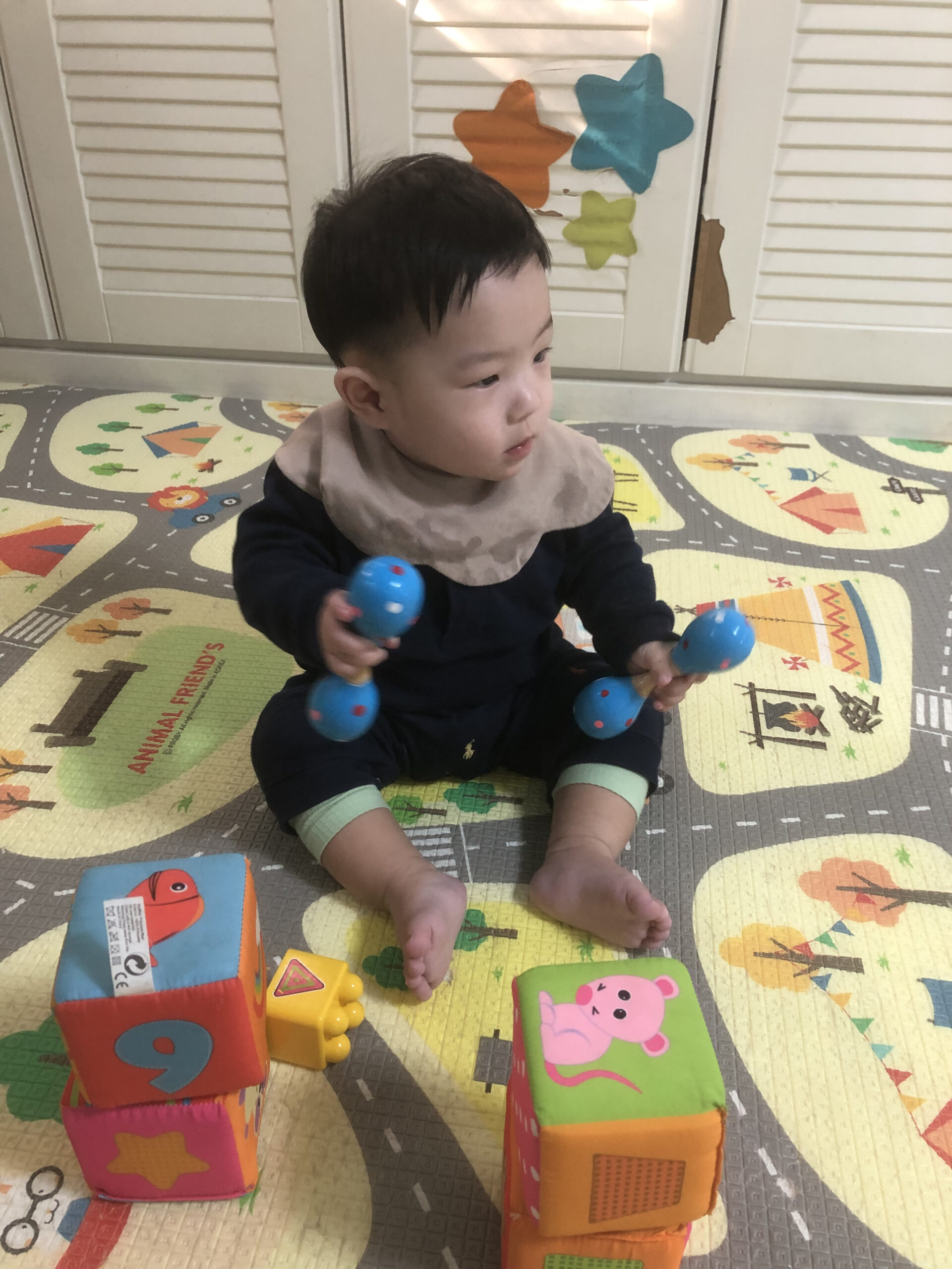 도로와 동물 패턴이 그려진 놀이매트 위에 앉아 파란색 딸랑이 두 개를 양손에 들고 옆을 바라보는 지호. 주변에 알록달록한 블록들이 놓여 있다.