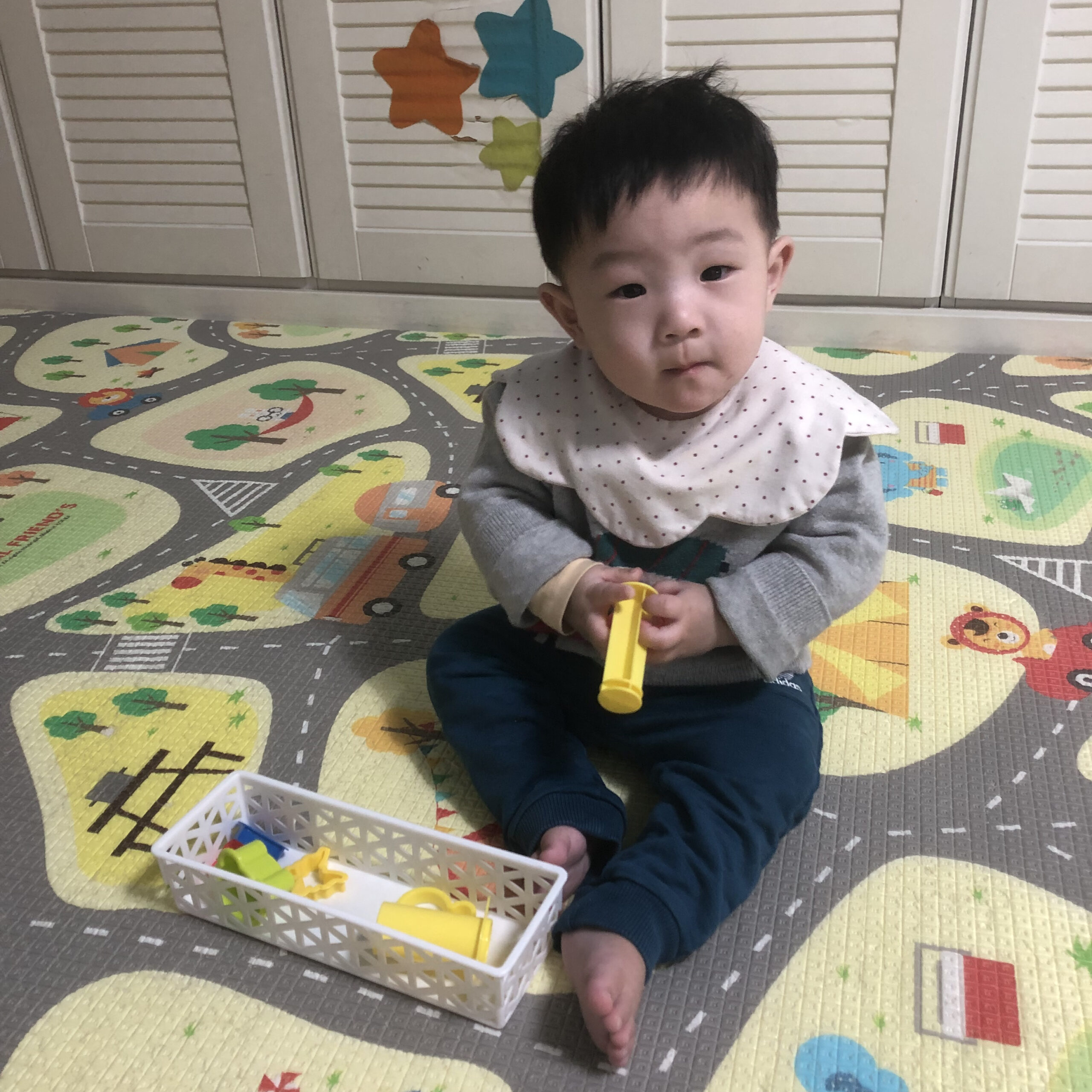 도로와 동물 패턴이 그려진 놀이매트 위에 앉아 노란색 장난감을 들고 카메라를 응시하는 지호. 옆에는 흰색 바구니에 장난감들이 담겨 있다.