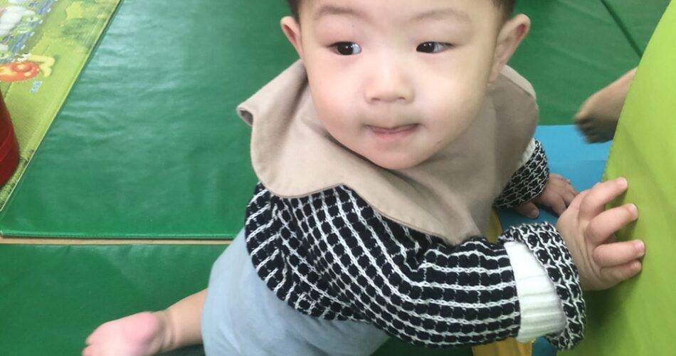 지호가 실내 놀이방의 초록색 매트 위에서 무릎을 꿇고 앉아 연두색 벽에 기대어 주변을 호기심 가득한 눈으로 바라보고 있다. 검은색 체크무늬 상의와 베이지색 턱받이를 하고 있다.