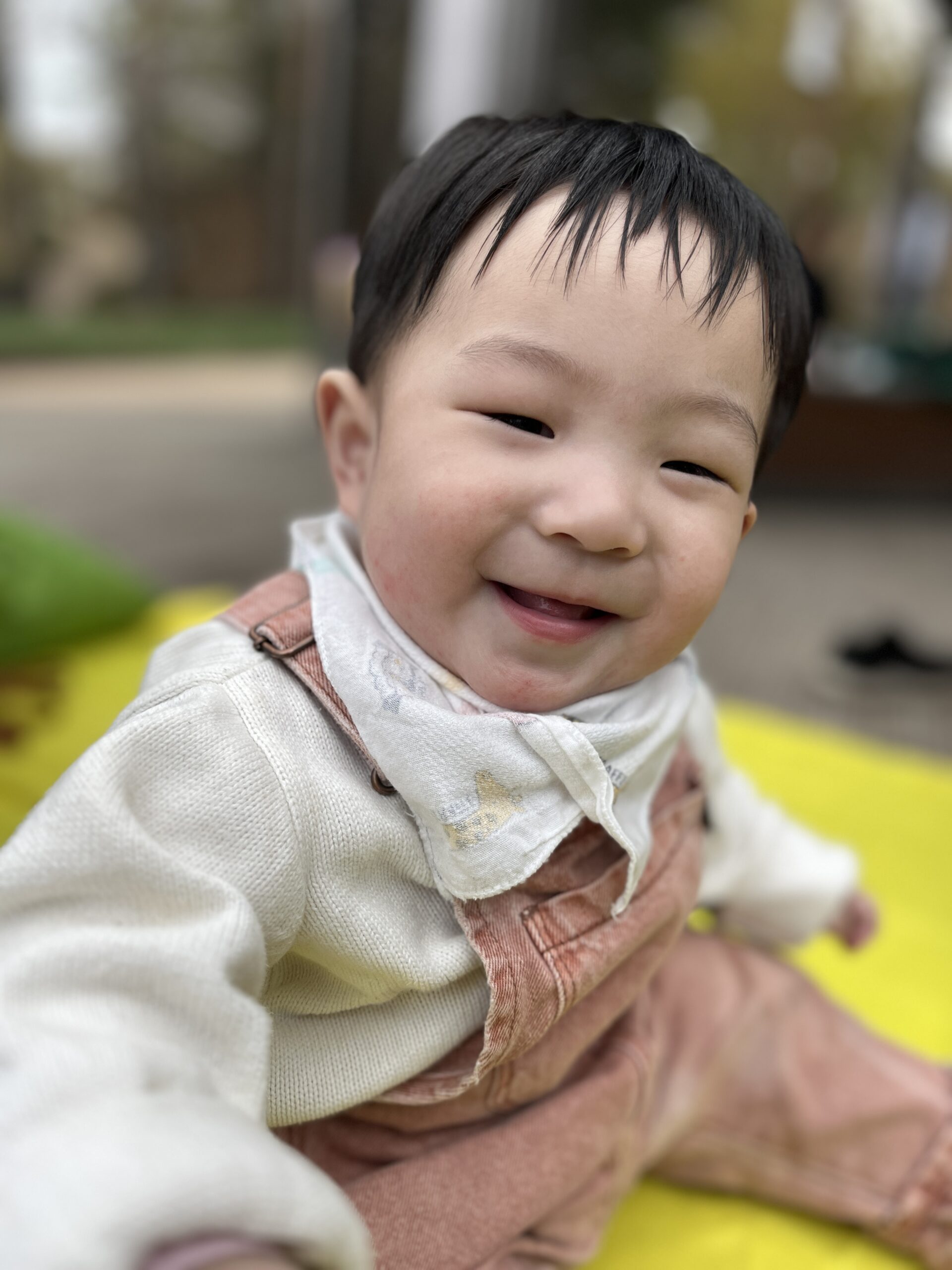 하얀 스웨터와 갈색 멜빵바지를 입고 흰색 턱받이를 한 지호가 환하게 웃으며 카메라를 바라보는 클로즈업 사진. 배경은 초록색과 노란색으로 흐려져 있다.