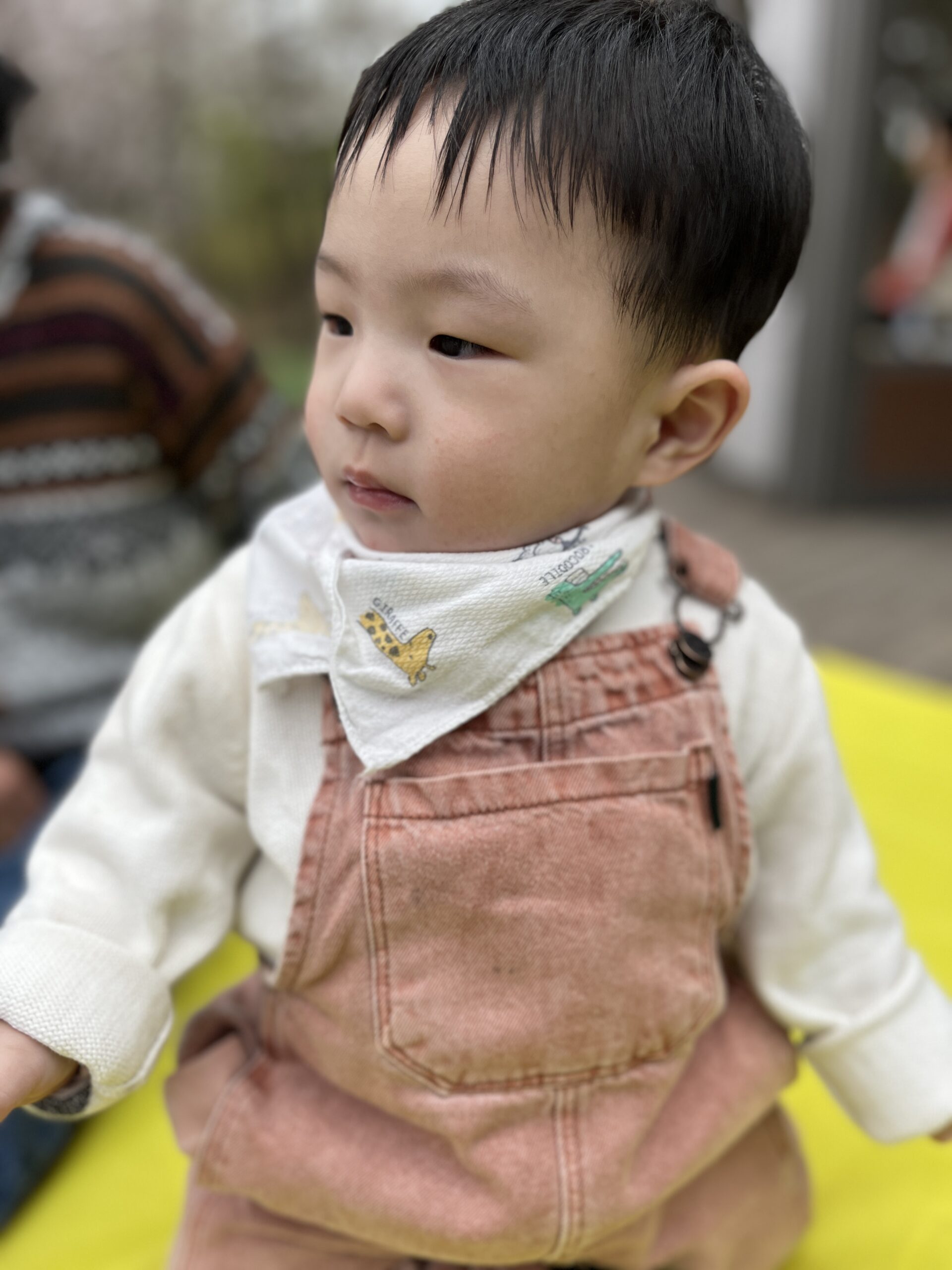 노란 매트 위에 앉아 베이지색 멜빵바지와 흰색 상의를 입고 목에는 패턴 턱받이를 한 아기 지호가 옆을 응시하고 있다. 배경은 흐릿한 야외 공간이다.