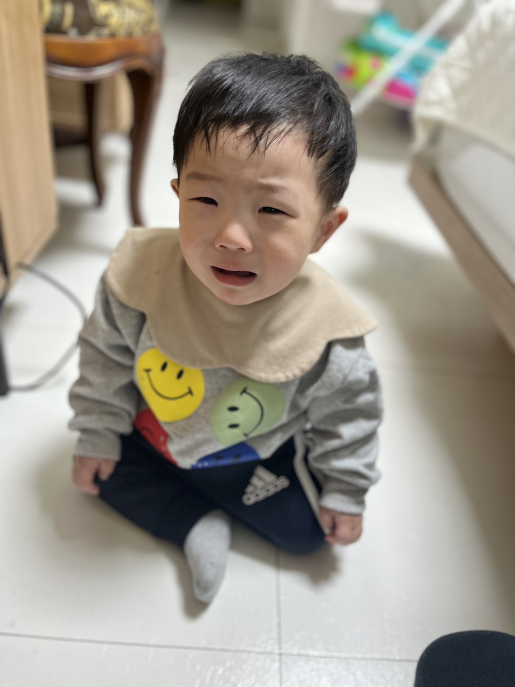 하얀 타일 바닥에 앉아있는 아기가 회색 스마일 얼굴 티셔츠와 검은색 바지, 그리고 베이지색 턱받이를 하고 있다. 울먹이는 표정으로 눈물을 그렁그렁 매단 채 카메라를 올려다보고 있다.