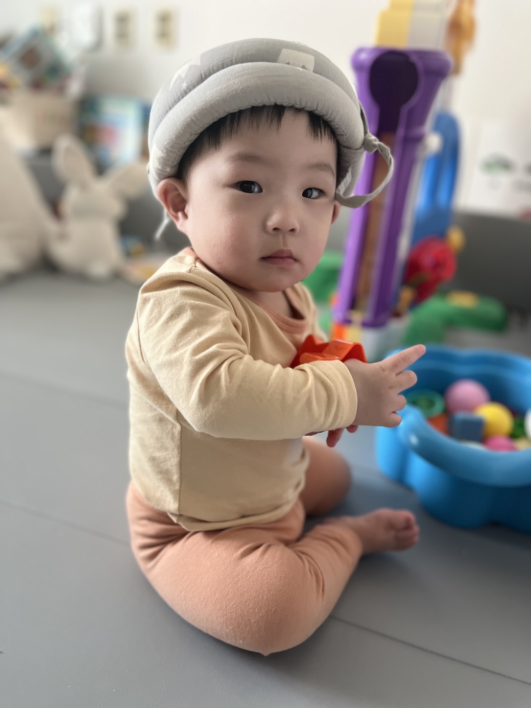 회색 안전 헬멧을 쓰고 연한 갈색 상의와 복숭아색 하의를 입은 아기 지호가 실내에서 바닥에 앉아 카메라를 보고 있다. 주변에는 알록달록한 장난감들이 놓여 있다.