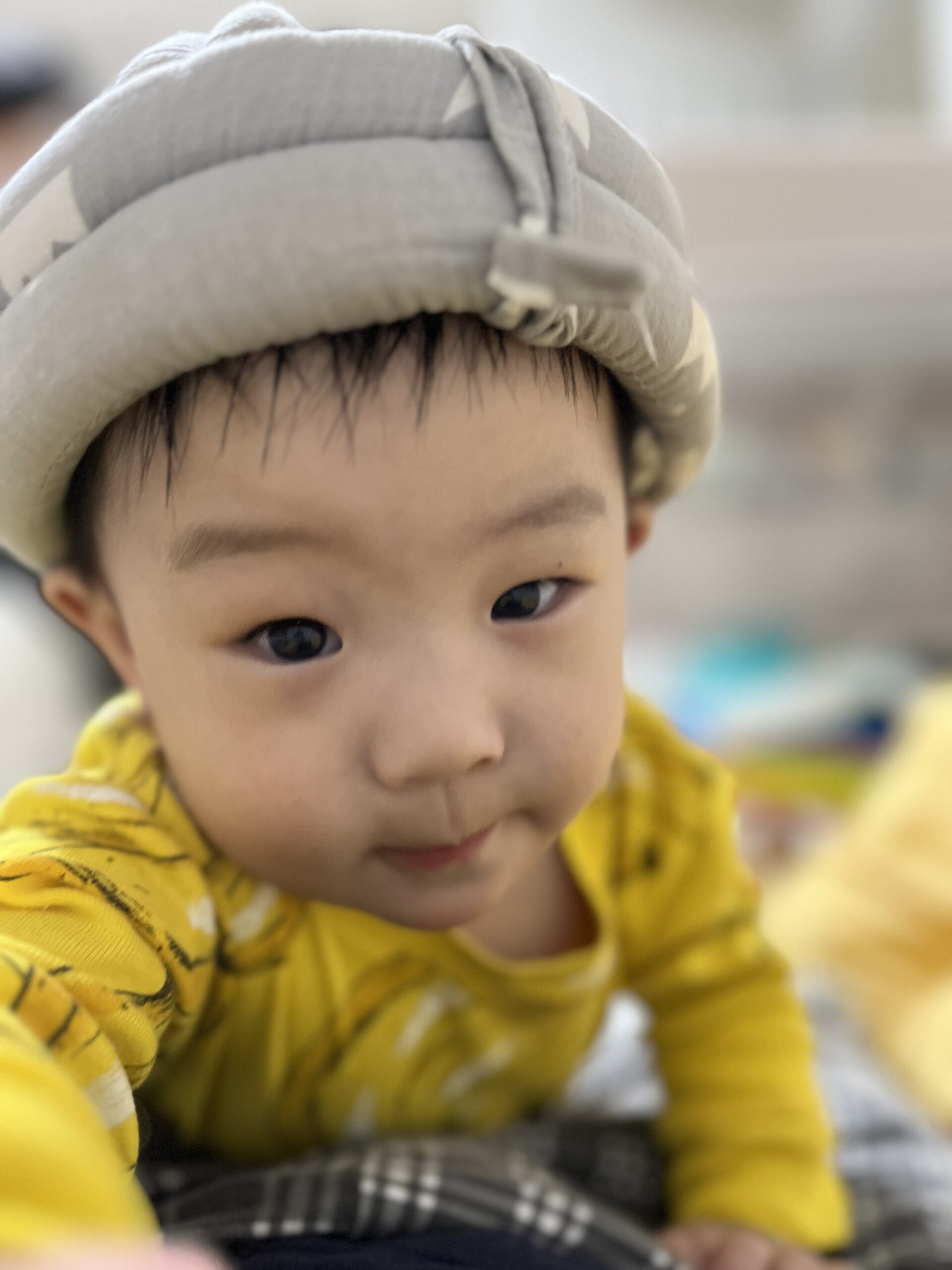 회색 보호 헬멧과 노란색 긴팔 티셔츠를 입은 아기의 얼굴이 클로즈업되어 있다. 바닥에 엎드린 자세로 카메라를 향해 호기심 가득한 눈빛으로 바라보고 있다.