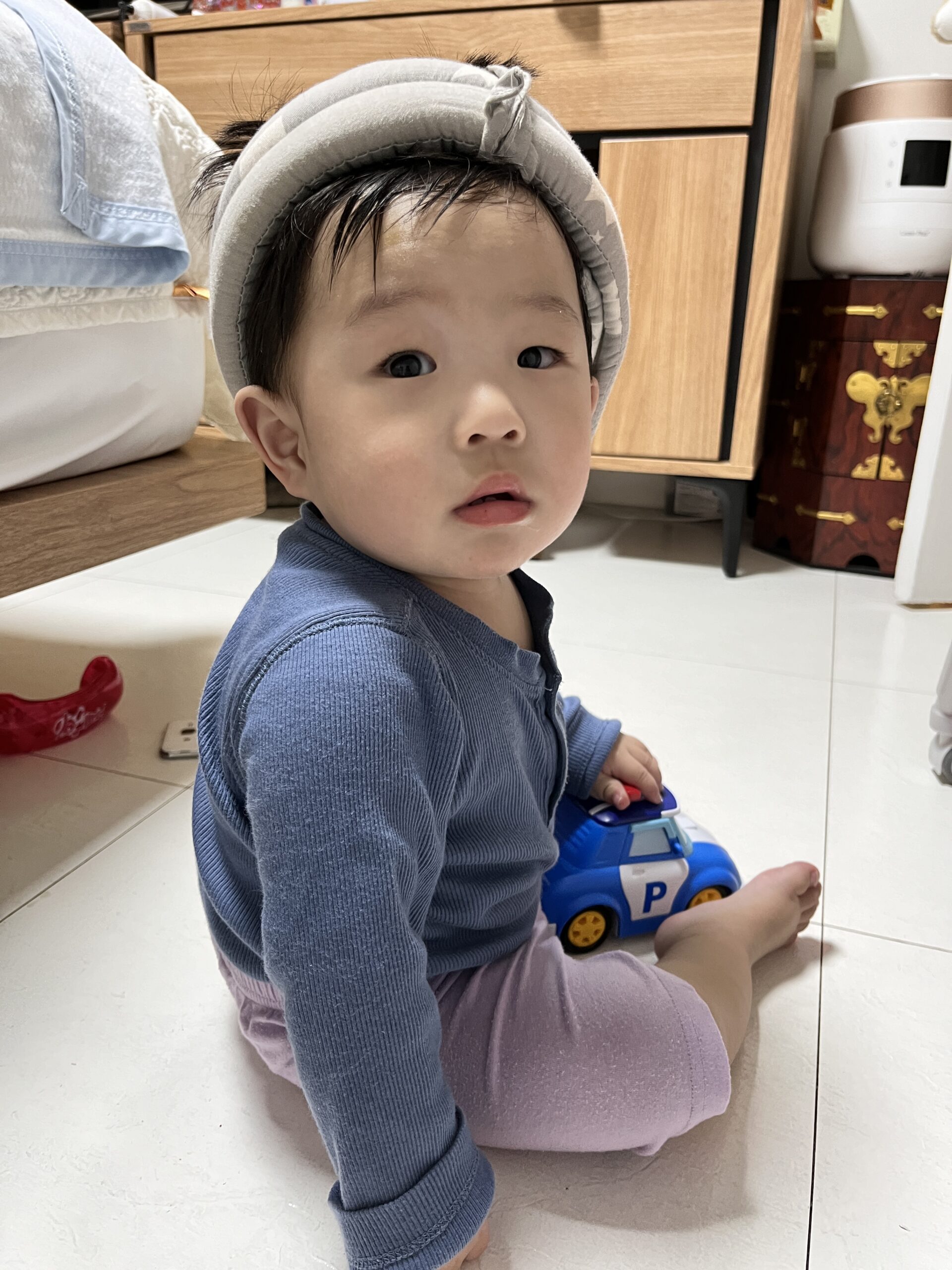 하얀 타일 바닥에 앉아있는 아기가 파란색 긴팔 티셔츠와 보라색 바지를 입고 회색 보호 헬멧을 쓰고 있다. 오른손에는 파란색 장난감 자동차를 쥐고 카메라를 진지하게 올려다보고 있다. 뒤로는 가구들이 보인다.