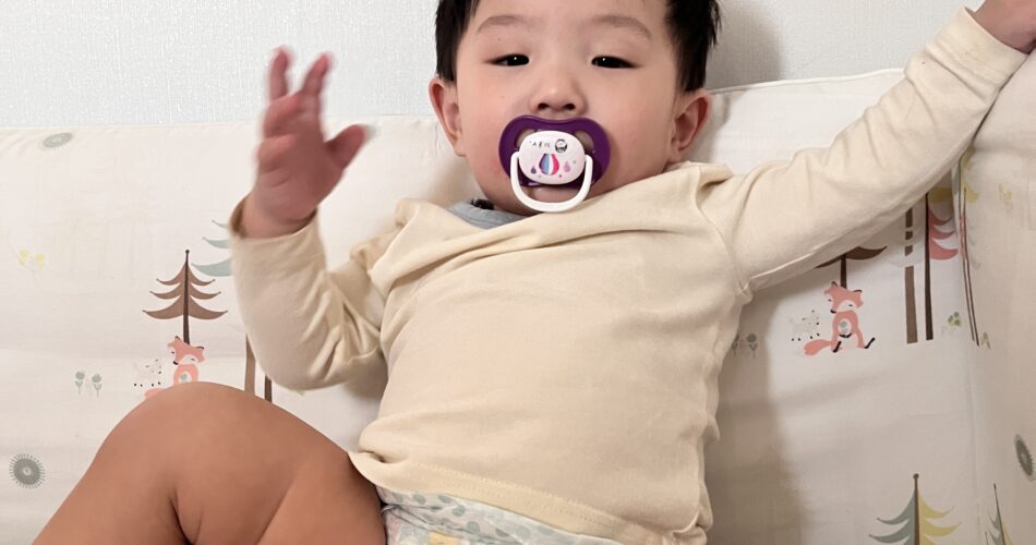 흰색 벽과 나무 패턴의 침대 가드에 기대어 앉아 있는 아기가 하늘색 상의와 기저귀만 입은 채 쪽쪽이를 물고 두 팔을 들어 만세를 하고 있다. 카메라를 바라보는 눈빛이 개구지다.