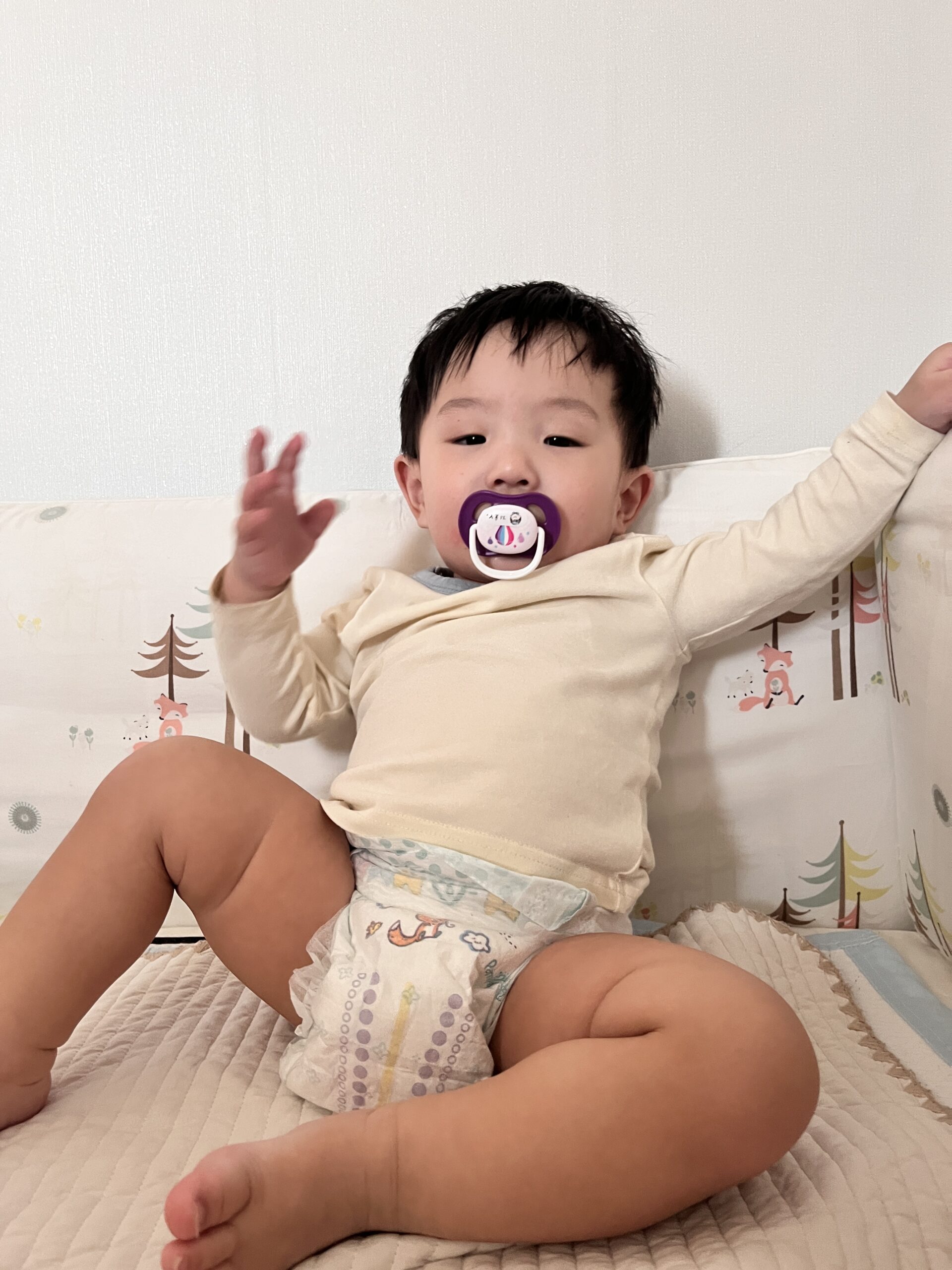흰색 벽과 나무 패턴의 침대 가드에 기대어 앉아 있는 아기가 하늘색 상의와 기저귀만 입은 채 쪽쪽이를 물고 두 팔을 들어 만세를 하고 있다. 카메라를 바라보는 눈빛이 개구지다.