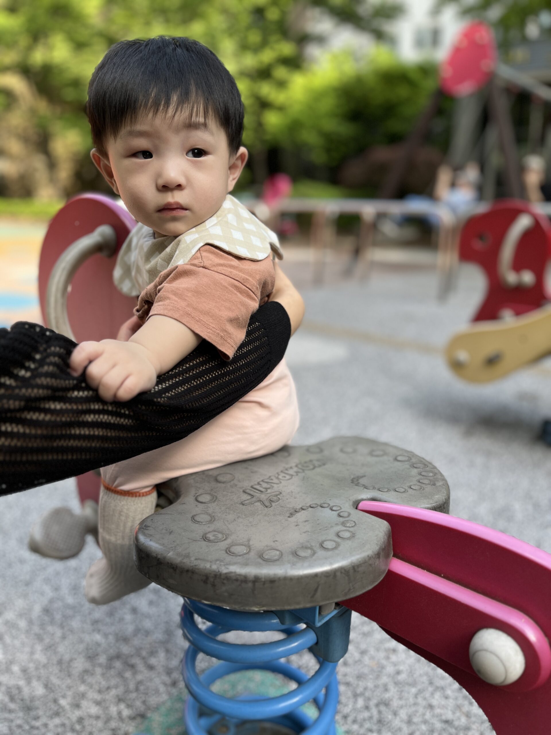 놀이터에서 어른의 팔에 의지해 스프링 놀이기구에 앉아 있는 지호. 베이지색 턱받이와 분홍색 옷을 입고 진지한 표정으로 어딘가를 바라보고 있다.