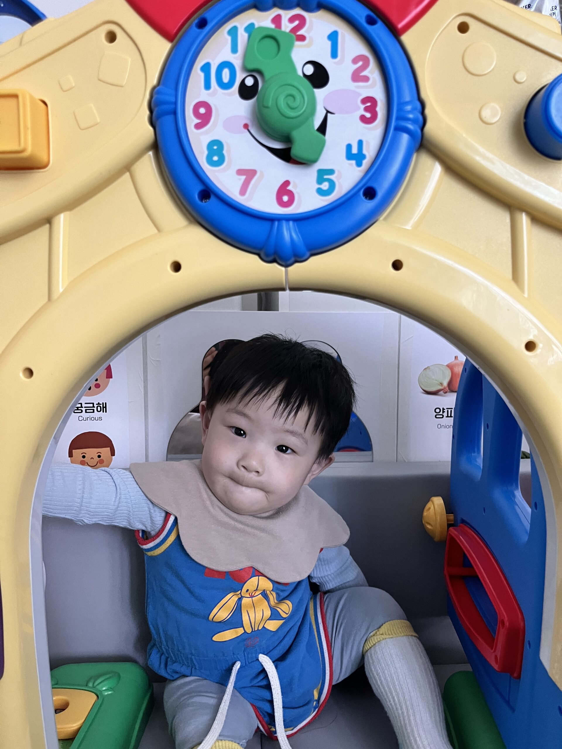 파란색 토끼 그림 상의와 갈색 턱받이를 한 아기 지호가 노란색 장난감 집 안에 앉아 입을 앙 다문 귀여운 표정으로 카메라를 바라보고 있다.