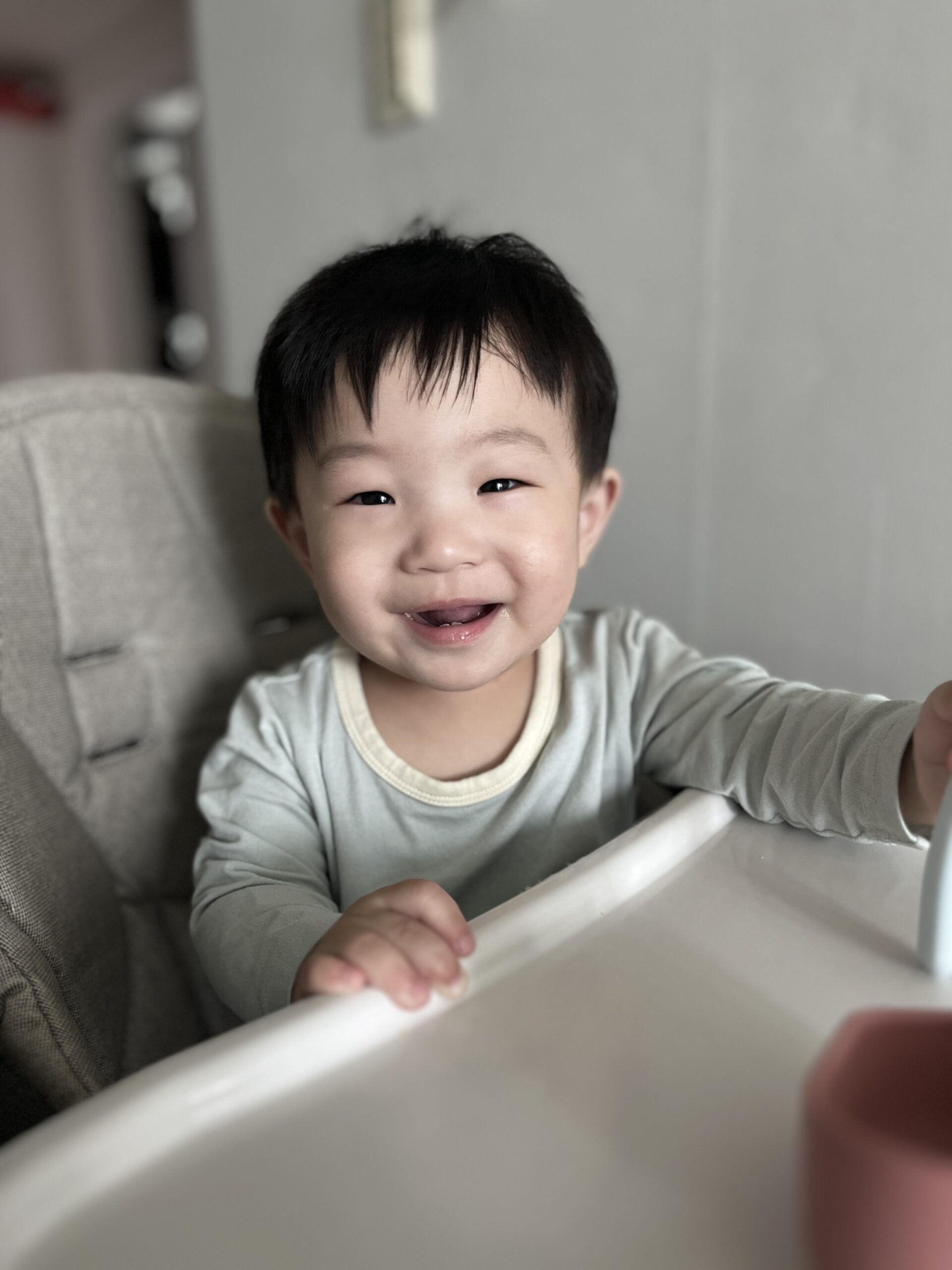 하이체어에 앉아 정면을 바라보며 환하게 웃고 있는 아기 지호. 연두색 긴팔 옷을 입고 양손으로 식탁을 잡고 있으며, 눈이 휘어지는 귀여운 미소를 짓고 있다.