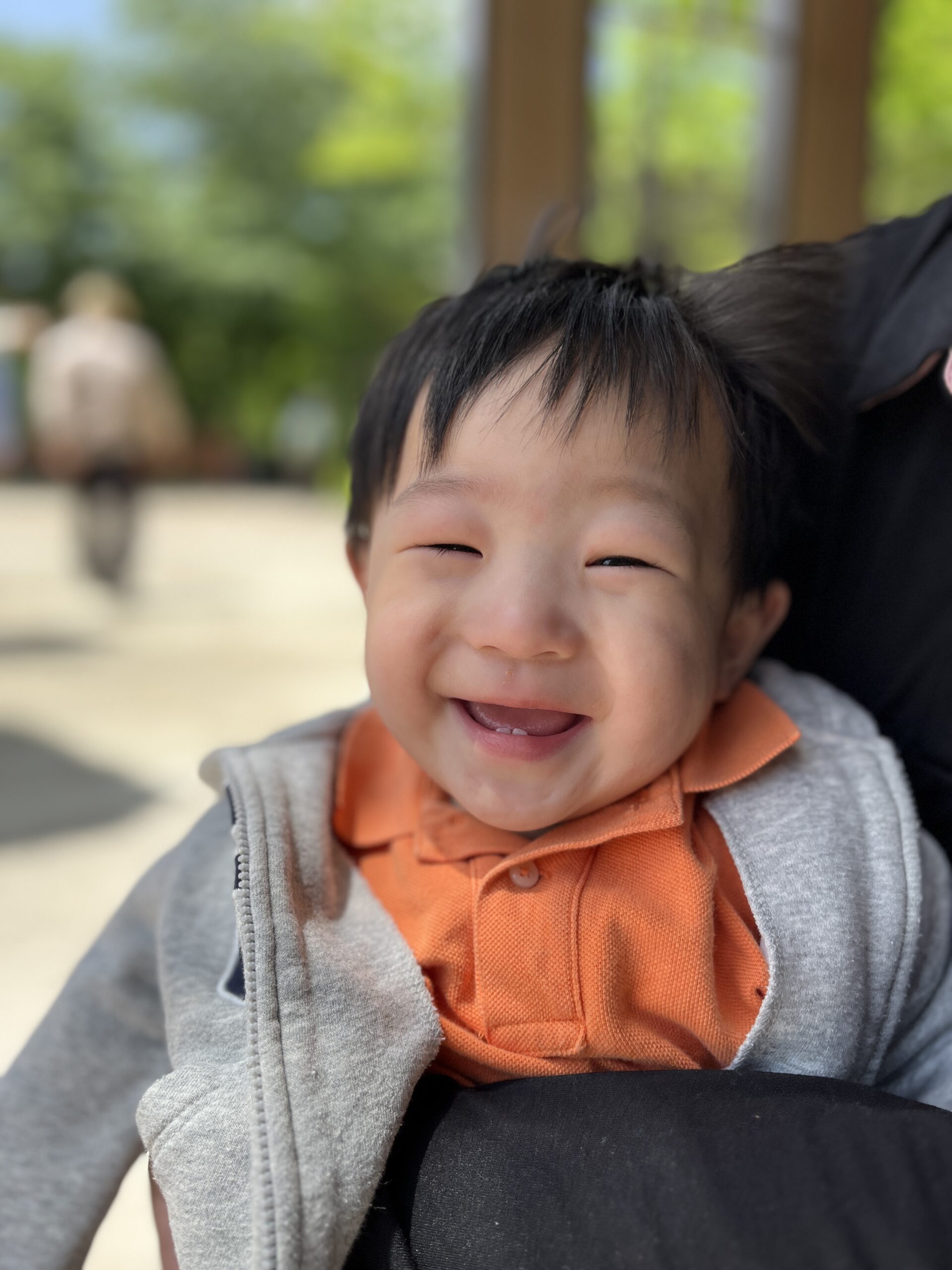 야외에서 햇살을 받으며 환하게 웃고 있는 아기 지호의 얼굴 근접 사진. 눈을 지그시 감고 활짝 웃고 있으며, 주황색 카라티 위에 회색 후드집업을 입고 있다. 배경은 밝게 흐려져 있다.