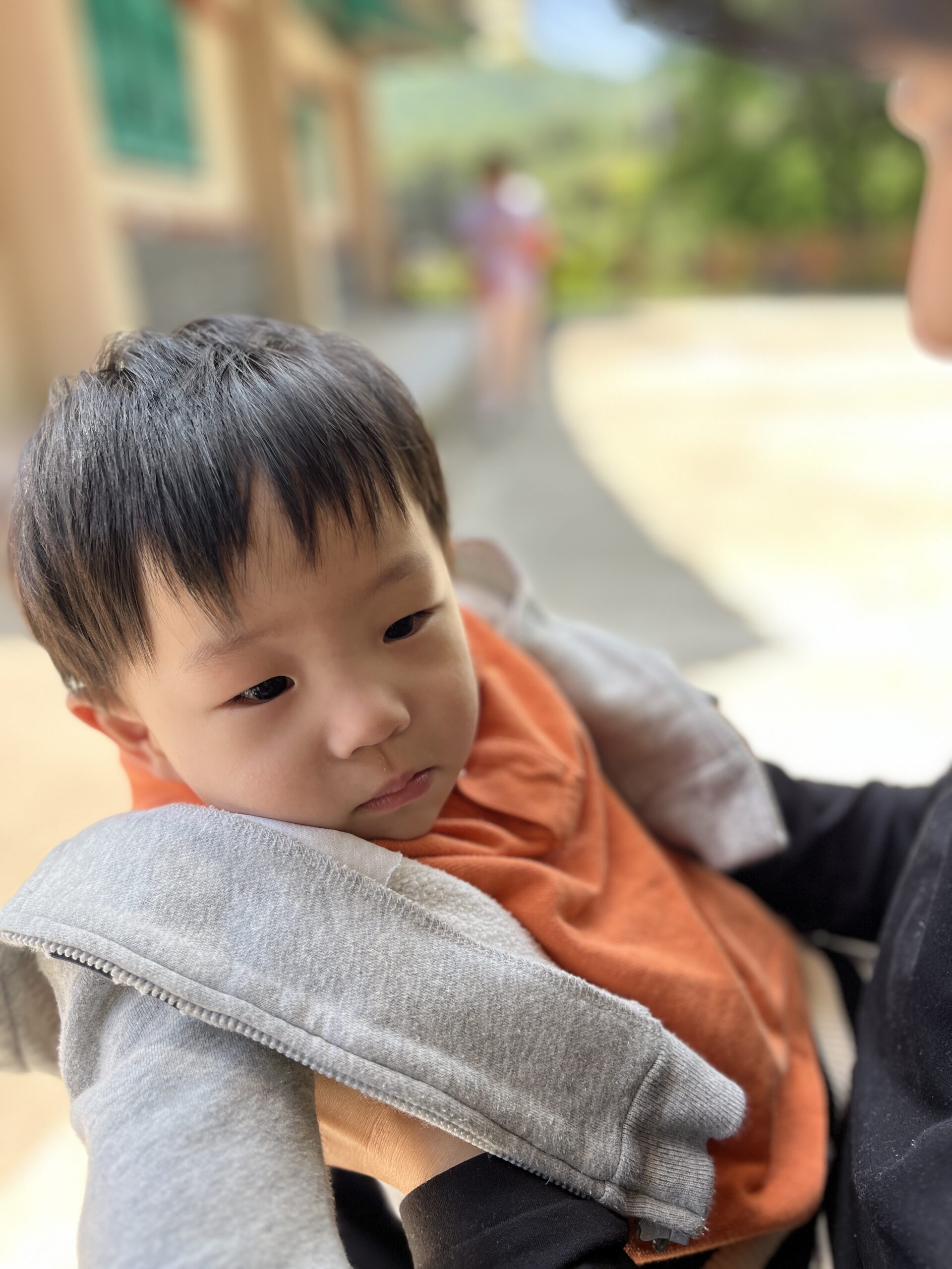 야외에서 어른의 품에 안겨 고개를 약간 숙이고 생각에 잠긴 듯한 표정을 짓고 있는 지호. 주황색 상의와 회색 후드집업을 입고 있다.