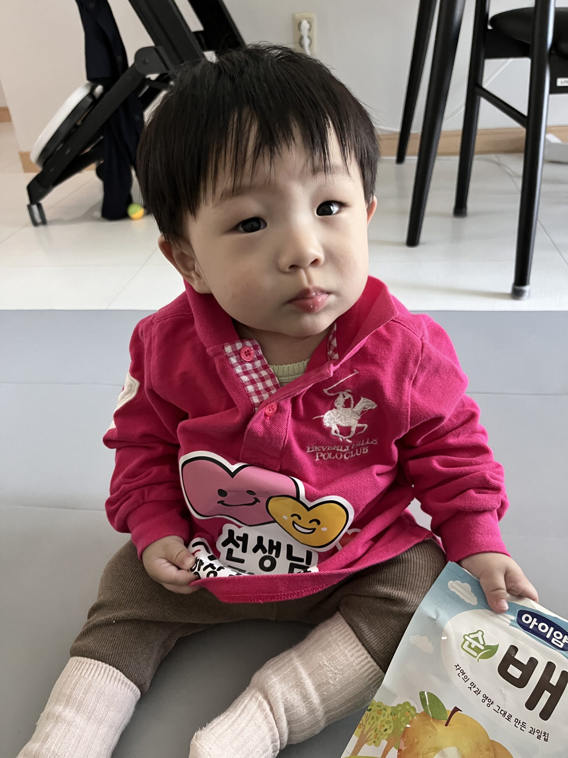 실내 회색 매트 위에 앉아 있는 지호. 분홍색 '선생님' 글자가 새겨진 상의와 갈색 하의를 입고, '아이얌 순 배' 아기 간식 파우치를 들고 카메라를 응시하고 있다.