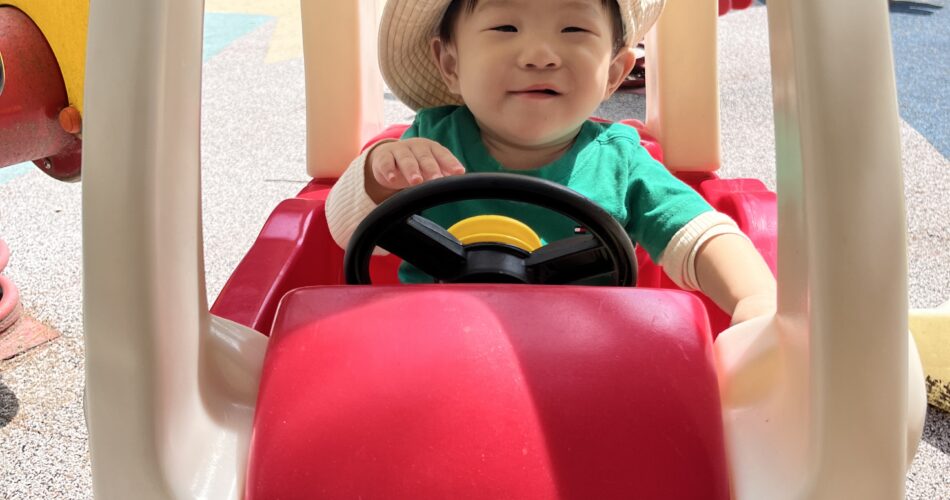 밝은 햇살 아래 놀이터에서 빨간색 아기 자동차에 앉아 즐겁게 웃고 있는 지호. 베이지색 밀짚모자와 초록색 상의를 입고 운전대를 잡고 있다.