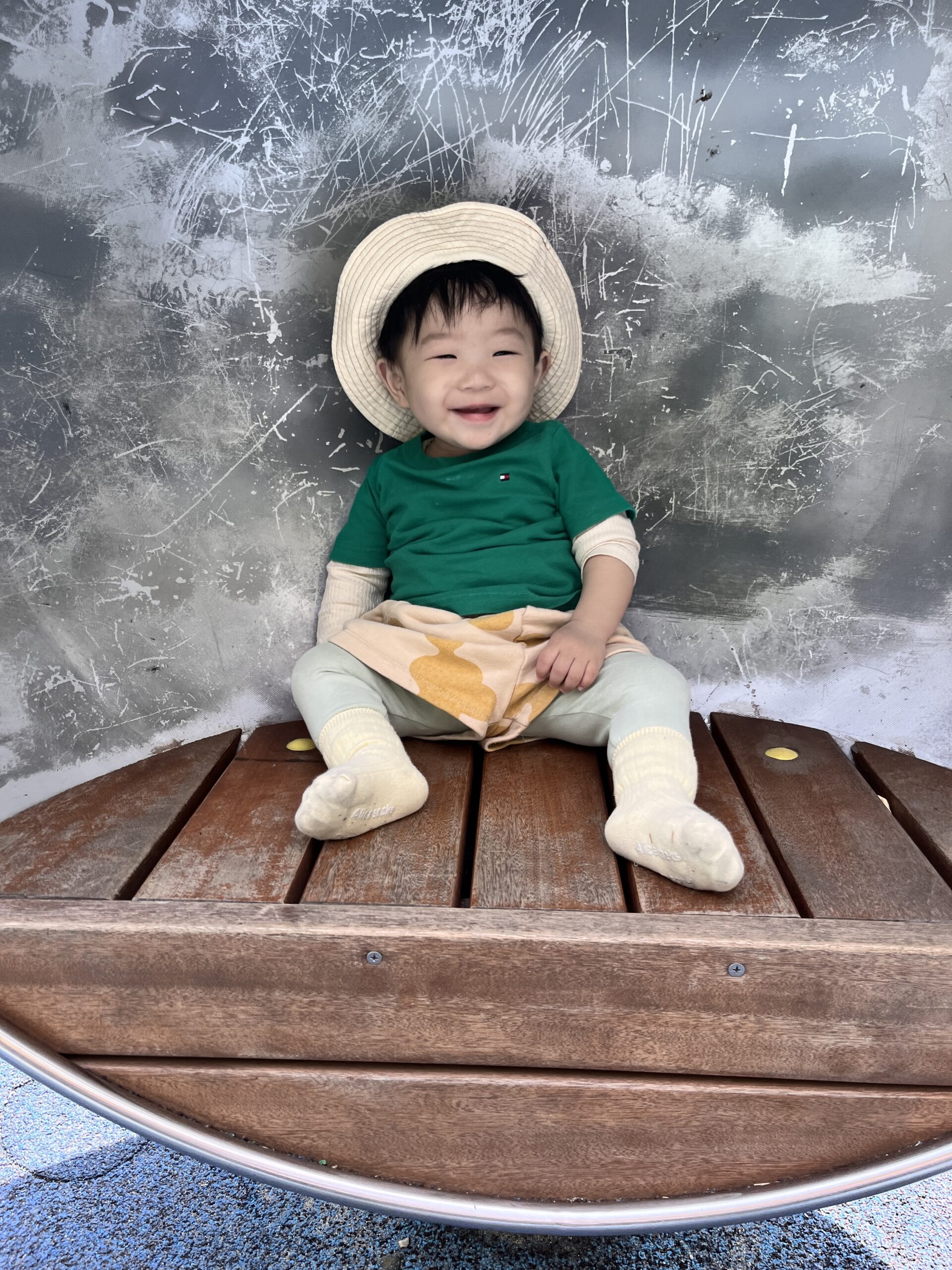놀이터의 나무 벤치 같은 곳에 앉아 환하게 웃고 있는 지호. 베이지색 밀짚모자와 초록색 상의를 입고 있다. 뒤로는 회색의 긁힌 듯한 벽면이 보인다.