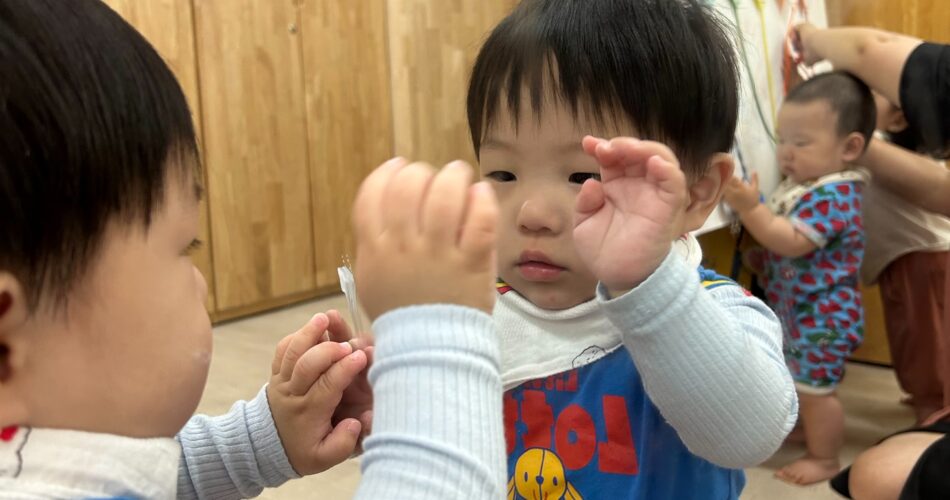 파란색 멜빵바지를 입은 아기 지호가 거울 속 자신을 신기한 듯 바라보며 한 손을 들어 올리고 있는 모습. 배경에는 나무 수납장과 다른 아기가 보인다.