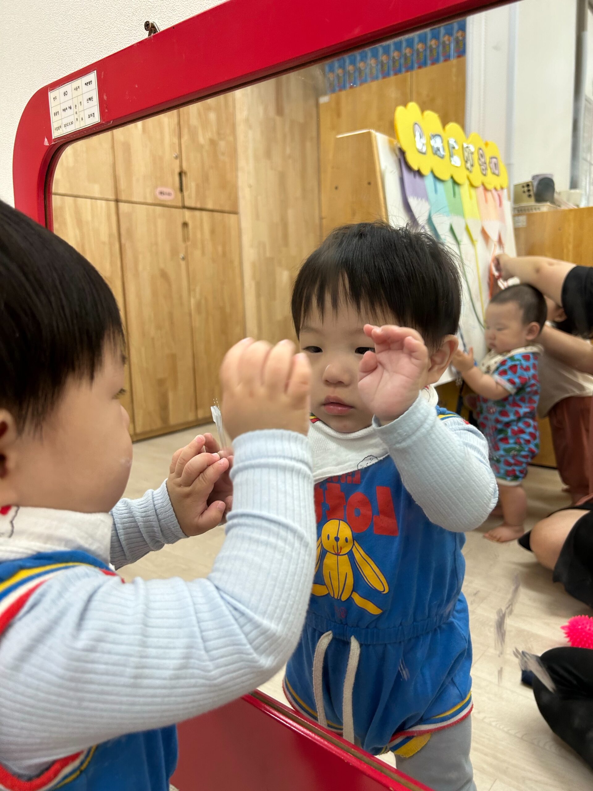 파란색 멜빵바지를 입은 아기 지호가 거울 속 자신을 신기한 듯 바라보며 한 손을 들어 올리고 있는 모습. 배경에는 나무 수납장과 다른 아기가 보인다.