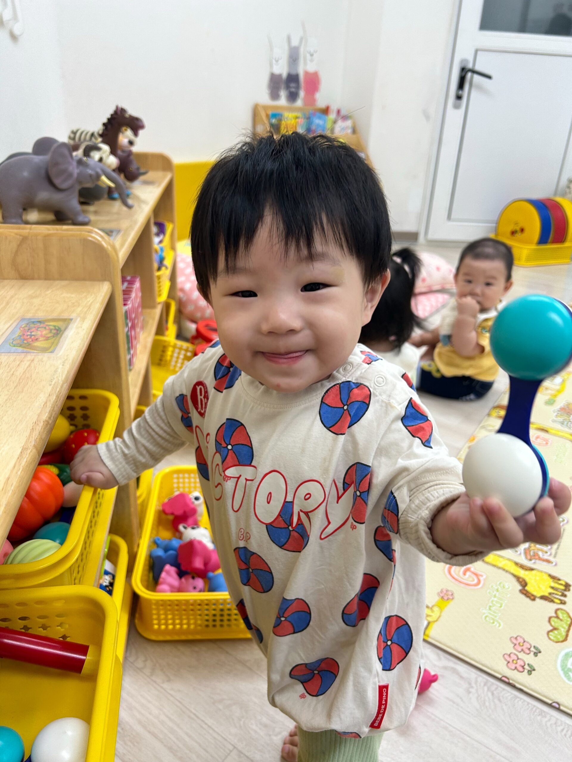 알록달록한 무늬의 상의를 입은 아기 지호가 장난감이 가득한 노란색 수납장 옆에서 환하게 웃으며 딸랑이를 들고 있는 모습. 뒤편에는 다른 아기가 보인다.