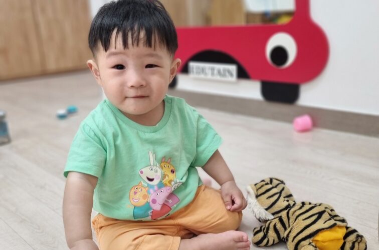 밝은 미소를 지으며 바닥에 앉아 있는 지호의 전신 사진. 연두색 티셔츠와 주황색 반바지를 입고 있으며, 뒤에는 빨간색 버스 모양 거울과 호랑이 인형, 그리고 여러 장난감들이 보인다.
