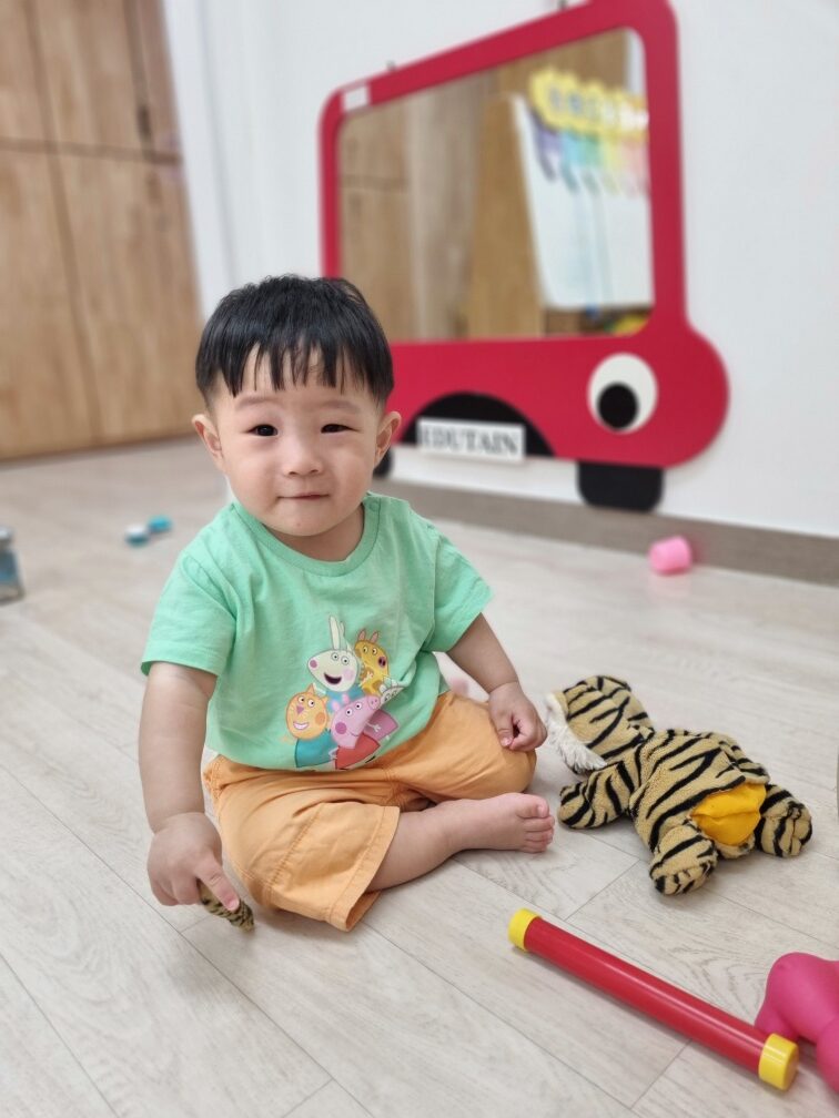 밝은 미소를 지으며 바닥에 앉아 있는 지호의 전신 사진. 연두색 티셔츠와 주황색 반바지를 입고 있으며, 뒤에는 빨간색 버스 모양 거울과 호랑이 인형, 그리고 여러 장난감들이 보인다.