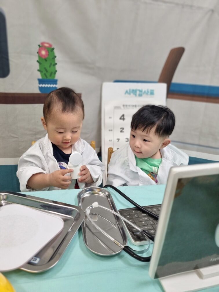 하얀 의사 가운을 입은 두 명의 아기가 테이블에 앉아 진지하게 놀이하는 모습. 지호는 오른쪽에서 진료 도구를 바라보고 있고, 다른 아기는 장난감을 들고 있다. 뒤편에 시력검사표가 보인다.